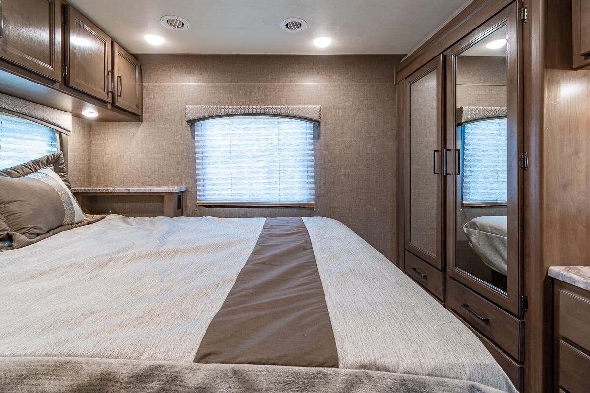 Comfortable RV rental bedroom featuring a bed, window with blinds, overhead cabinets, and a mirrored closet.. Thor Motor Coach Chateau 2020