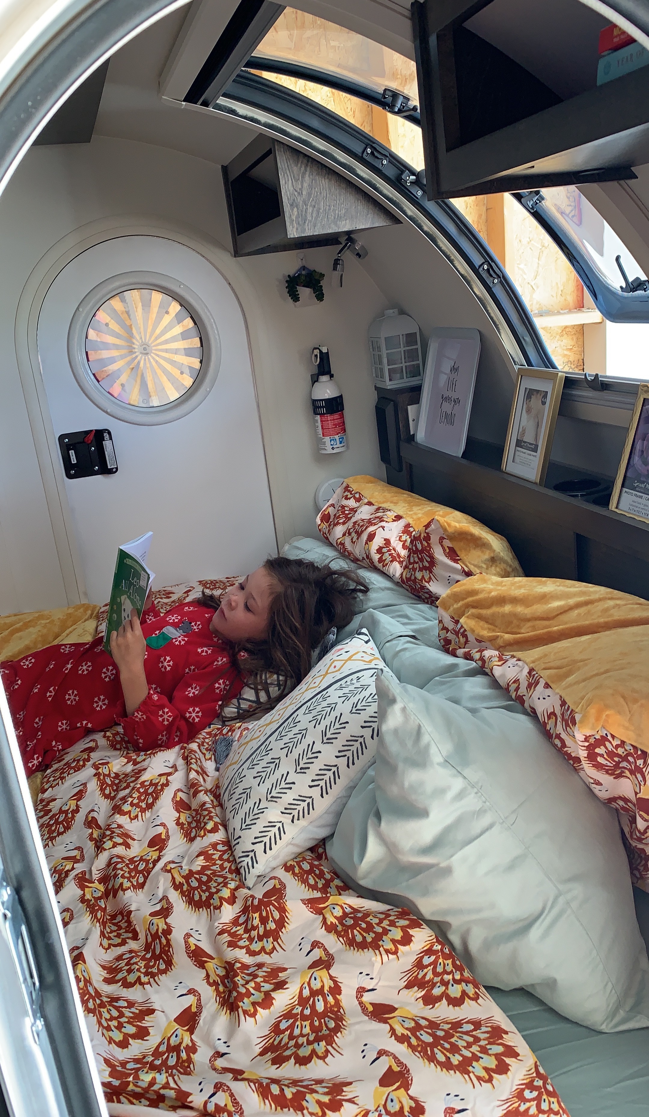 A child reads cozily in bed inside an RV rental. Peacock bedding, a round window, and a compact interior are visible.. T@G Txl 2020