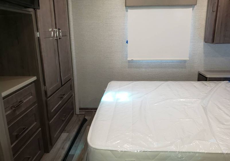 Compact RV rental bedroom featuring a new mattress (still wrapped), window shade, and wood-toned built-in storage.. Winnebago Minnie Winnie 2022