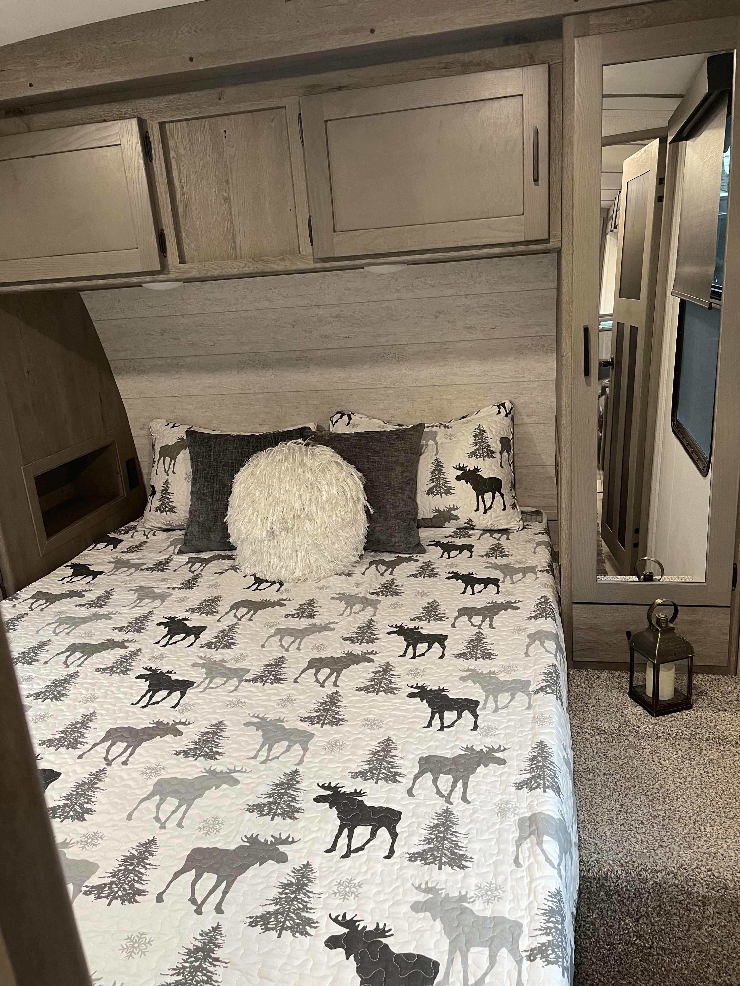 An RV rental bedroom featuring a bed with a moose patterned quilt, overhead cabinets, and a tall mirror on the right.. Keystone Outback 340BH 2021