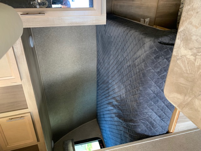 An RV rental interior features a bed covered in a dark blue quilted blanket, surrounded by light wood cabinetry and countertops.. Coachmen Catalina 2019