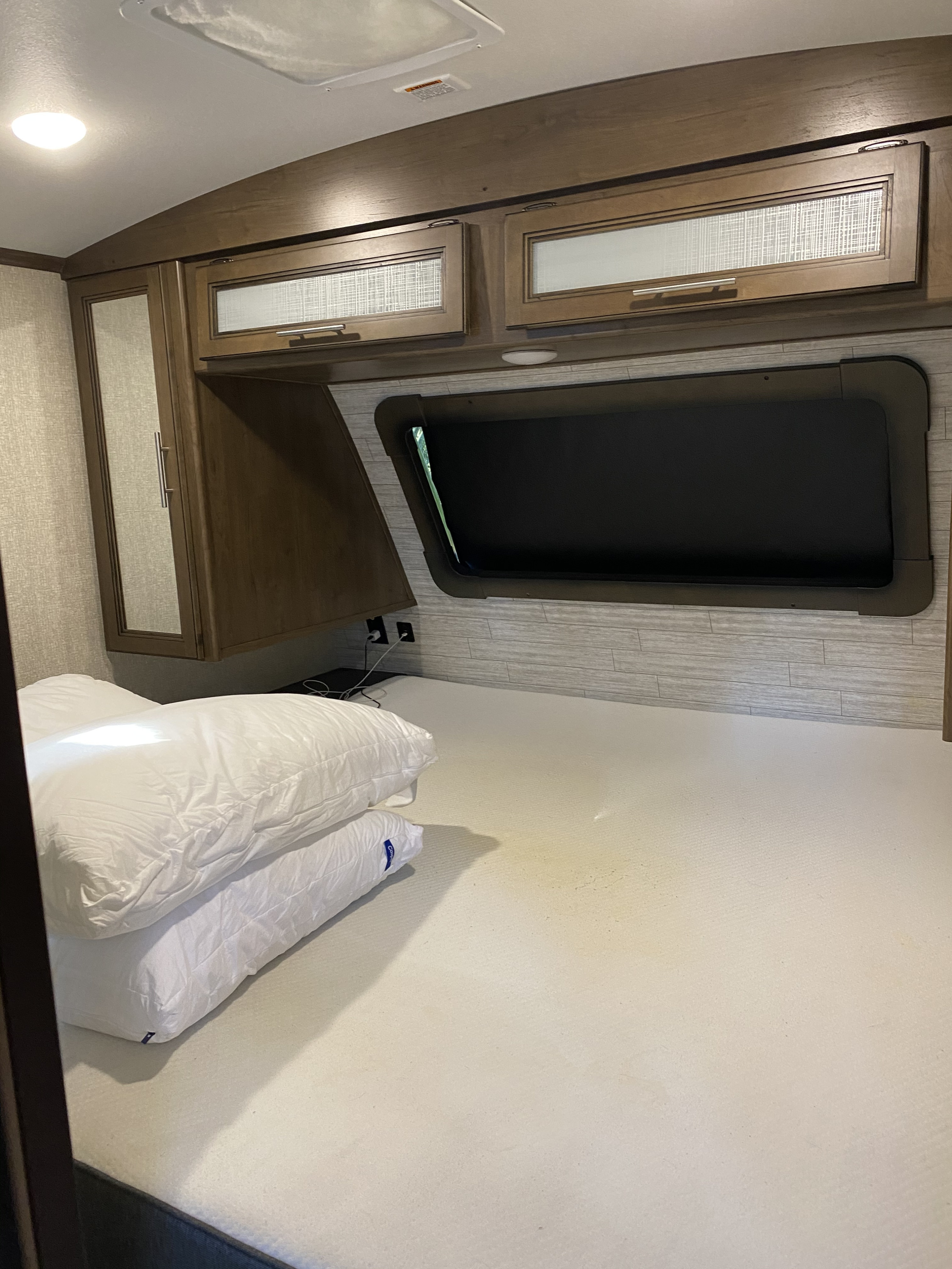 Inside an RV rental, a bedroom features a large bed, pillows, overhead cabinets, and a window with a dark shade.. Keystone Cougar Half-Ton 2020