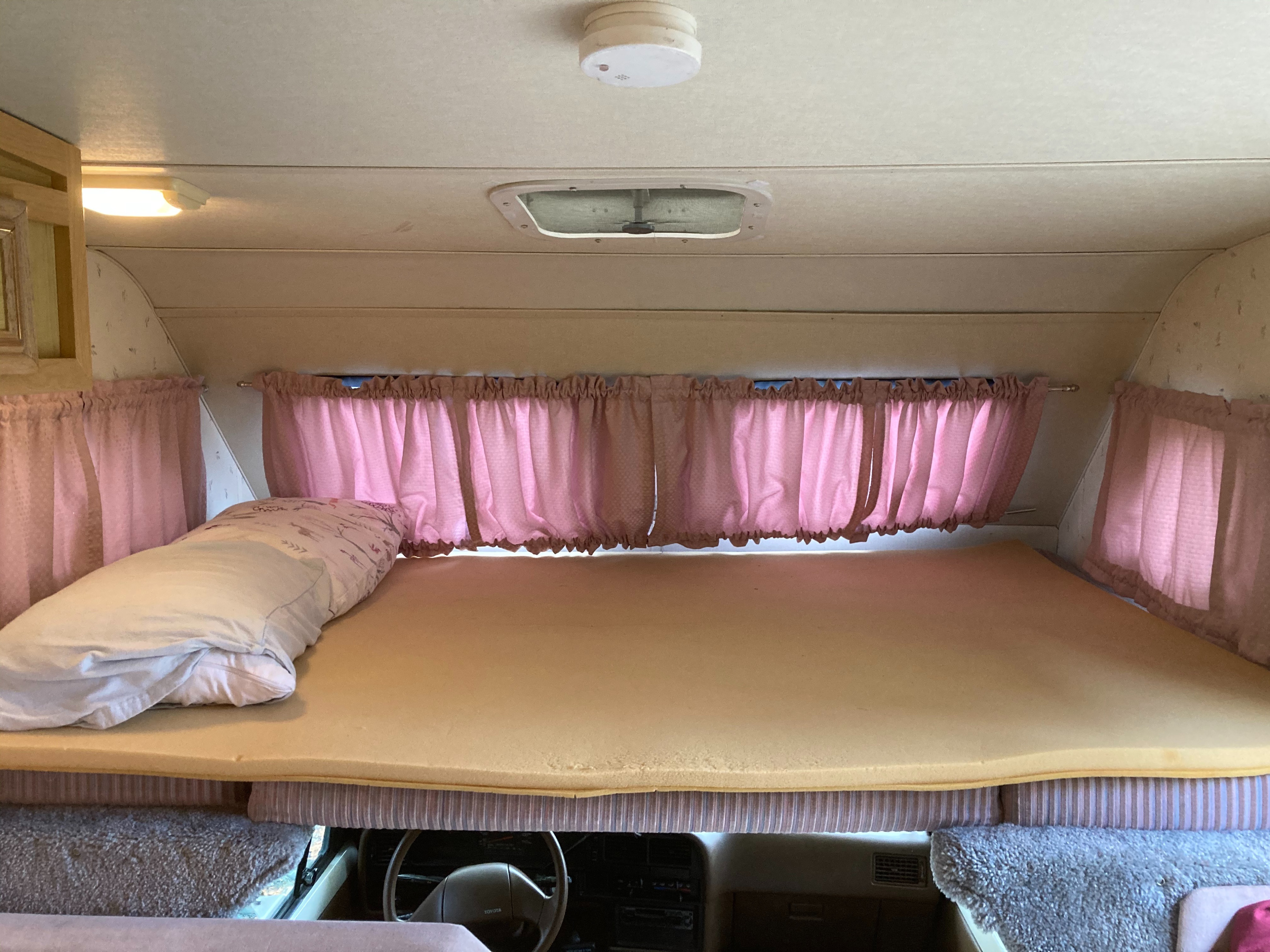 Cozy RV rental overhead bunk bed with foam mattress, pillow, and pink curtains, ideal for comfortable sleep on the road.. Winnebago Warrior 1991