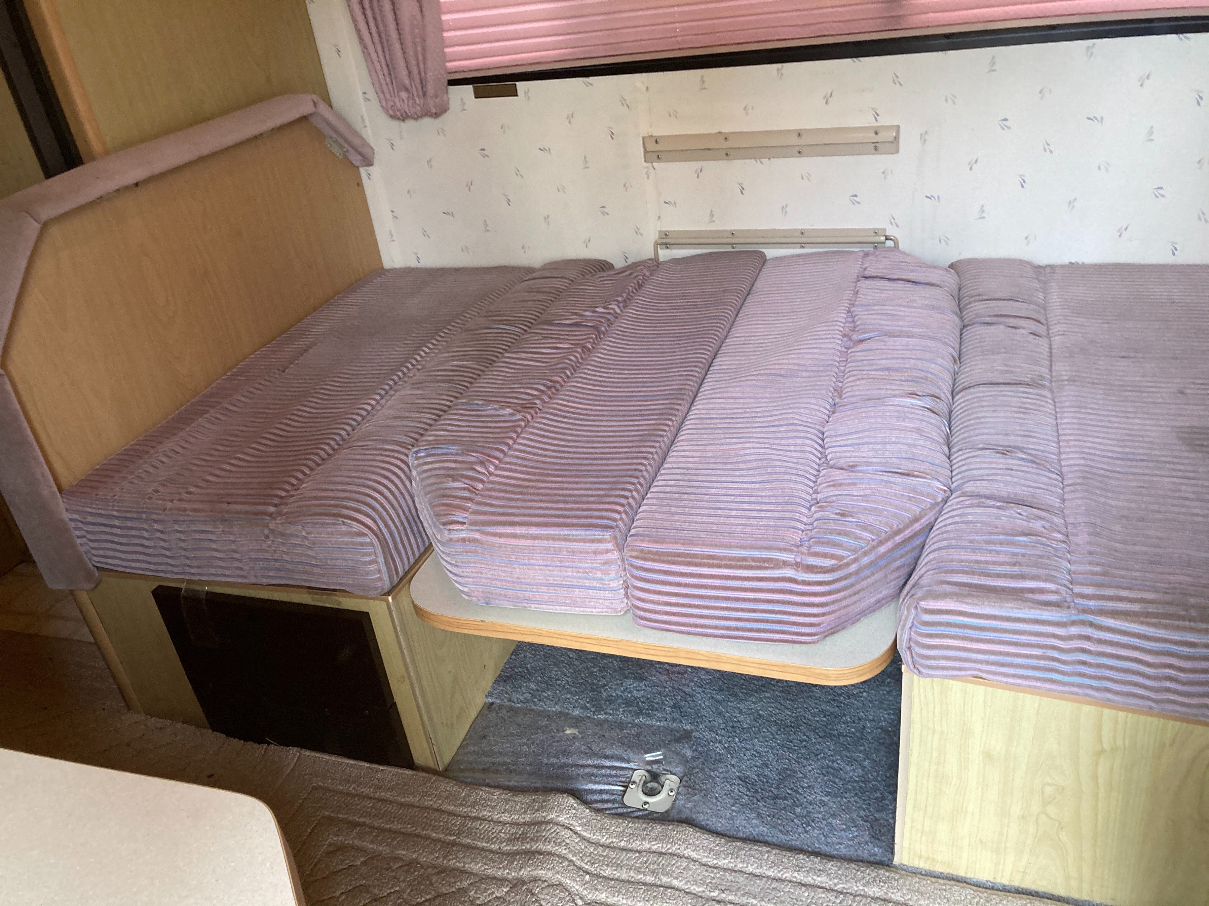 Inside an RV rental: a flexible bed and seating area with striped cushions, wooden cabinetry, and carpeted floor.. Winnebago Warrior 1991