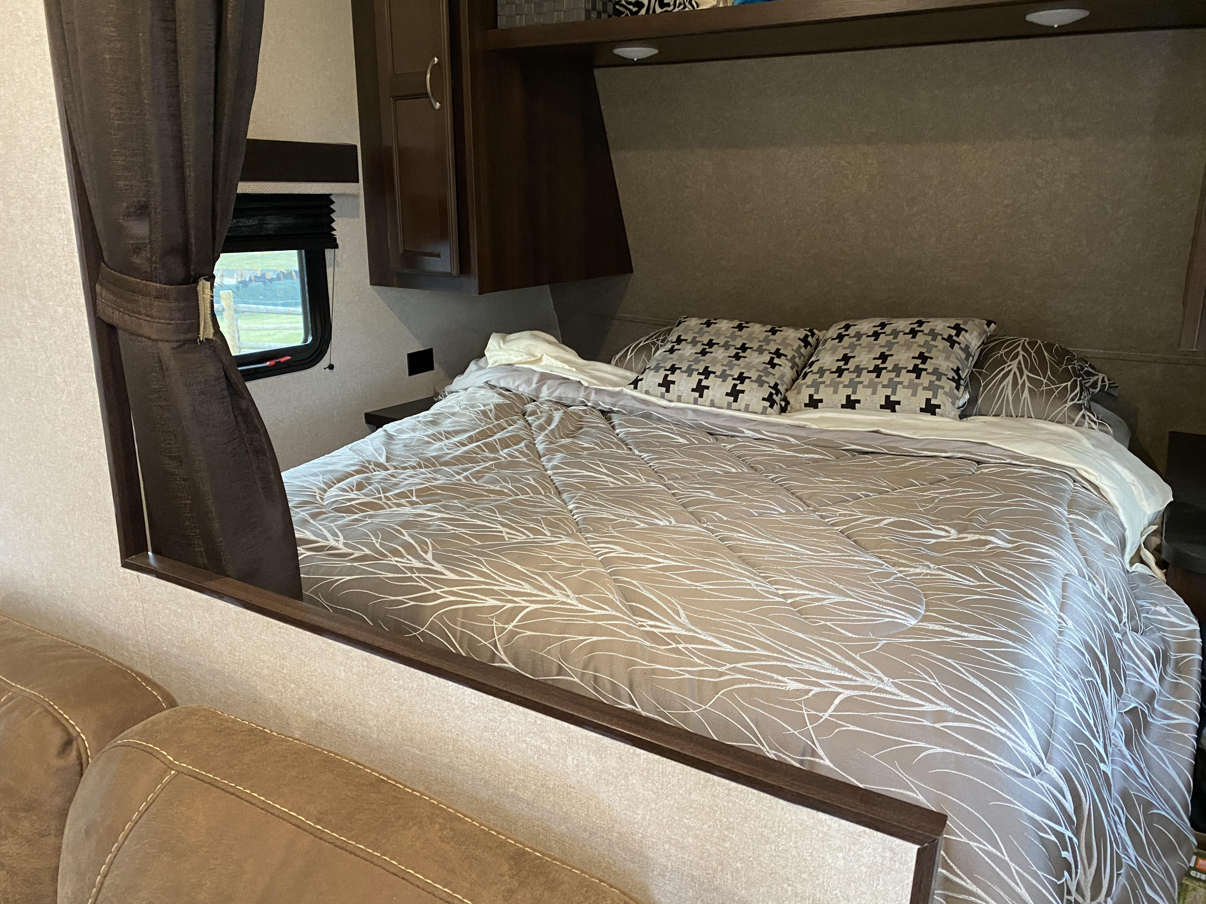 Cozy RV rental bedroom. Features patterned bedding, dark cabinets, window view, and foreground sofa.. Jayco Jay Flight 2018