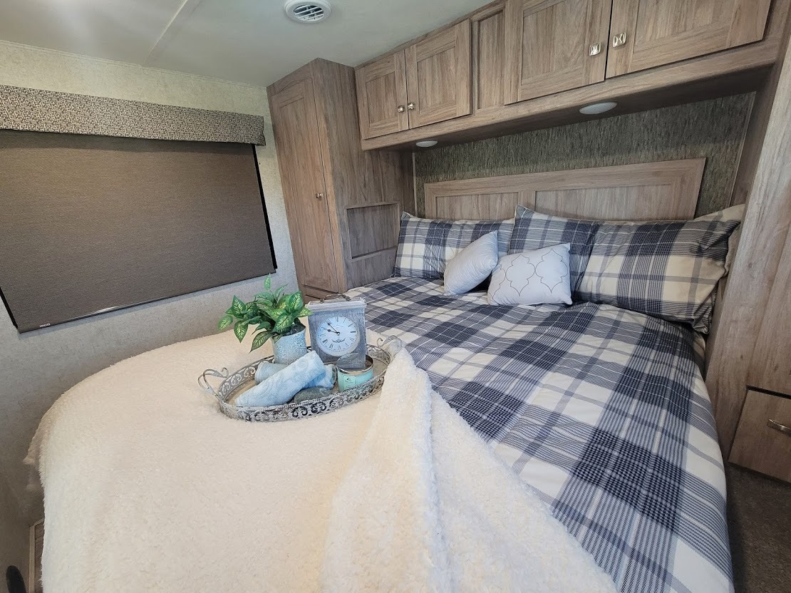 Cozy RV rental bedroom featuring blue and white plaid bedding, overhead storage cabinets, and a window with a roller shade.. Forest River Sunseeker 2019