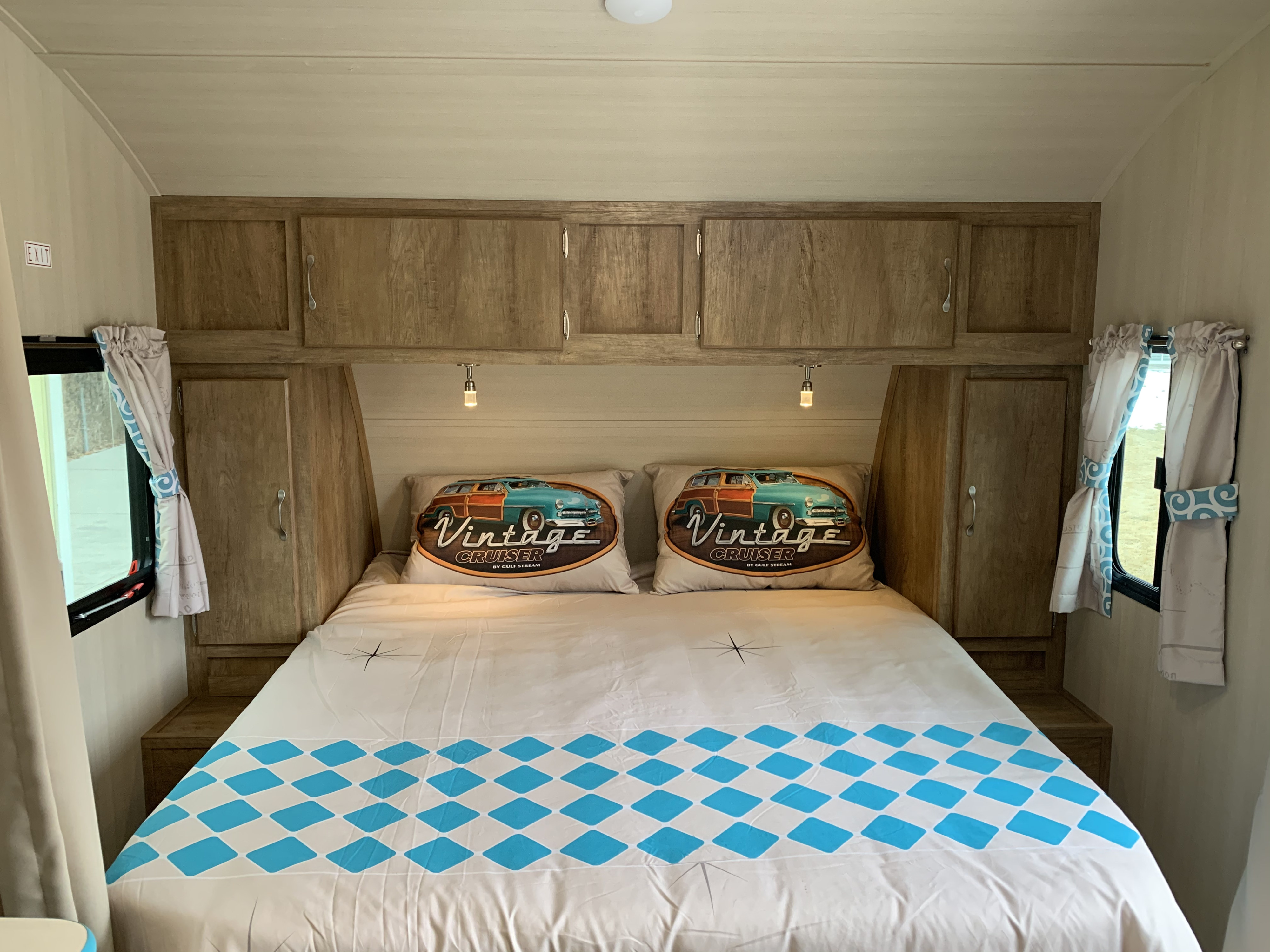 Inside a clean rv rental bedroom: a comfy bed with "Vintage Cruiser" pillows, wood cabinets, and side windows.. Gulf Stream Vista Cruiser 2021