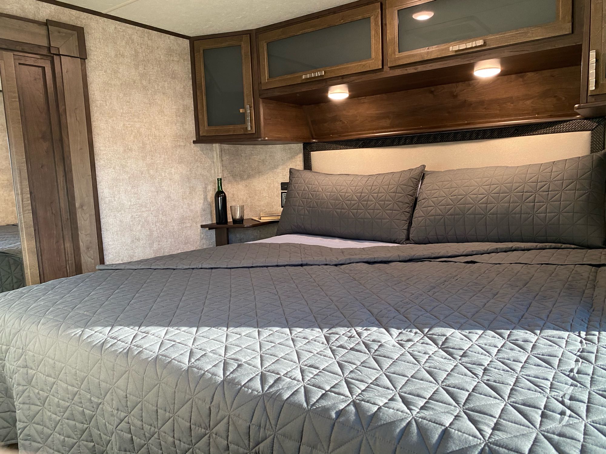 Inviting RV rental bedroom with a large bed, gray quilted bedding, and wooden overhead cabinets. A wine bottle is on the side shelf.. Open Range Highlander 2018