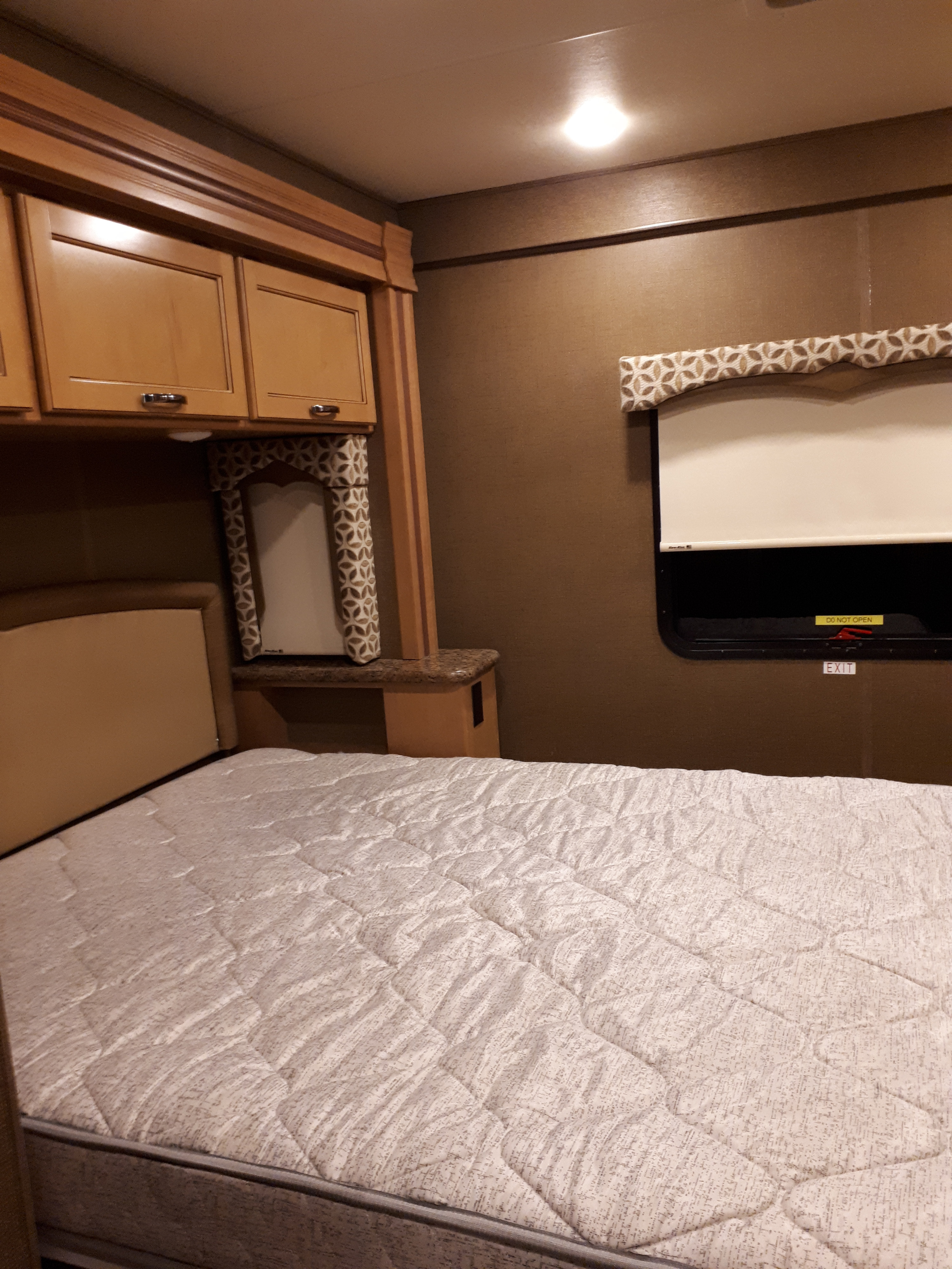 Inside an RV rental bedroom: a bed with a quilted mattress, wooden overhead cabinets, and a window with a shade and emergency exit sign.. Thor Four Winds 2017