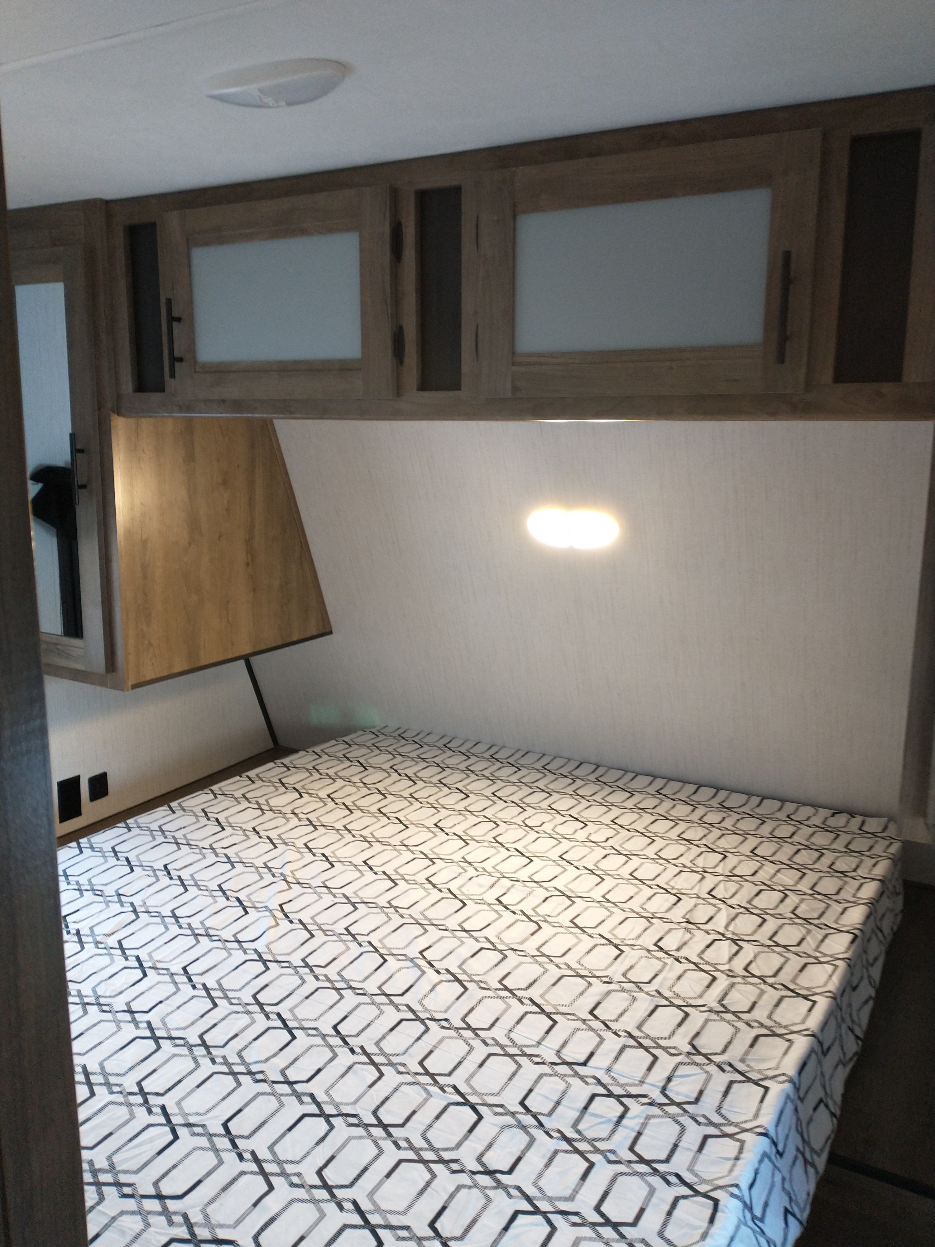 RV rental bedroom with a geometric bedspread, overhead storage cabinets, and a wall-mounted light.. Heartland Pioneer 2021