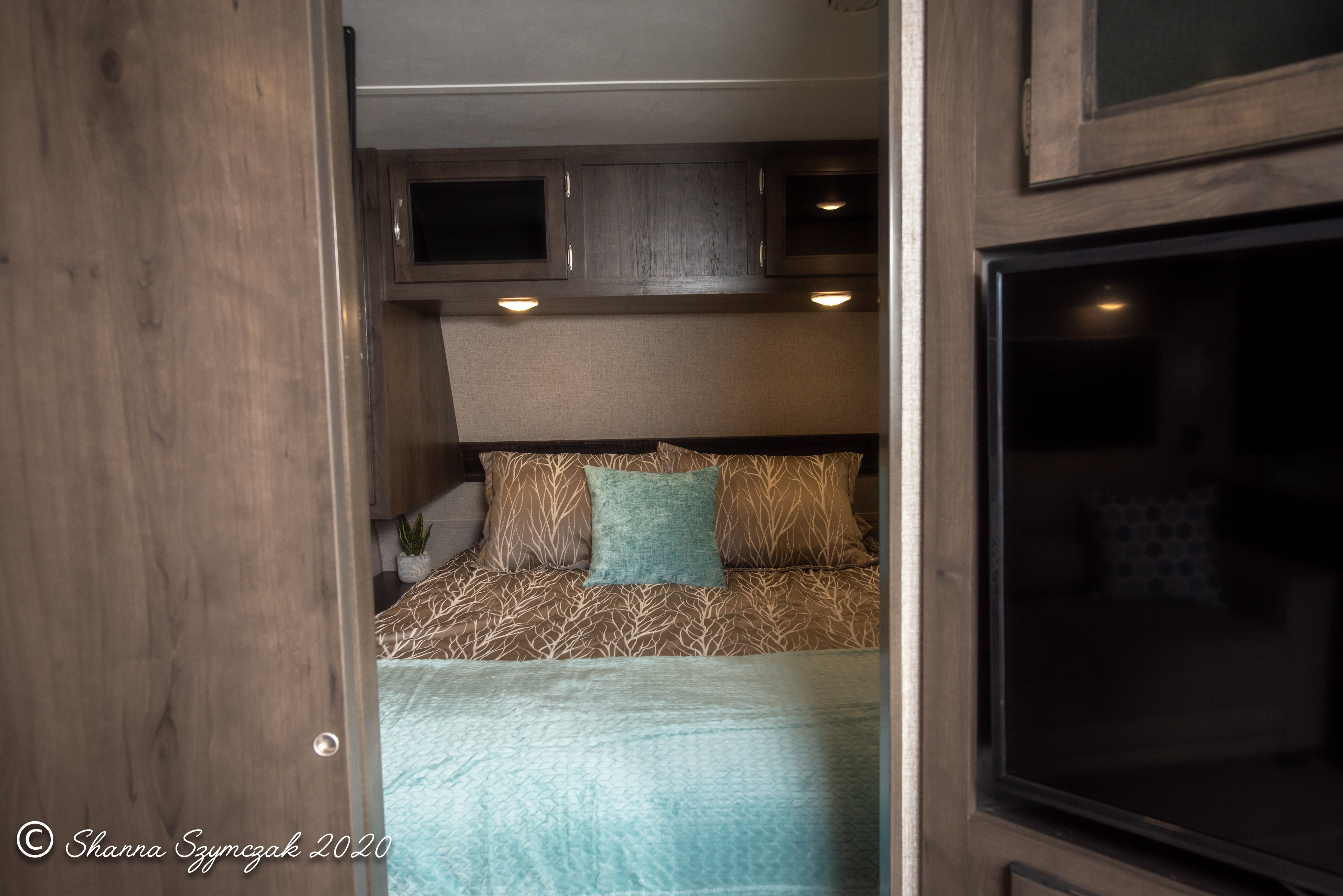Inside an RV rental: a compact, comfy bedroom with brown & teal bedding, overhead storage, and soft lights.. Jayco Jay Flight 2021