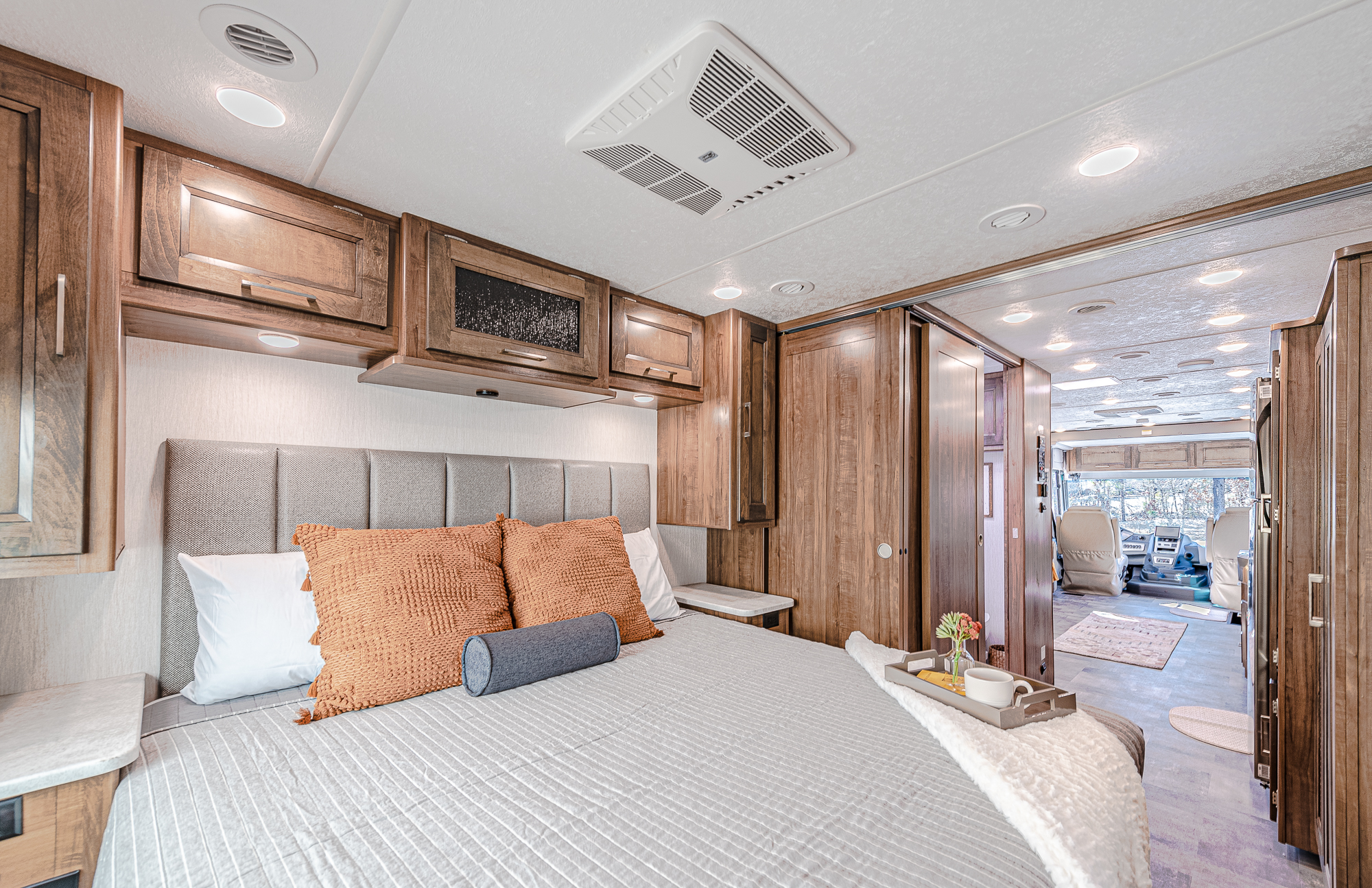 Inside a cozy RV rental: bedroom with patterned headboard, bright pillows, and warm wood accents, opening to front living area.. Coachmen Mirada 2021