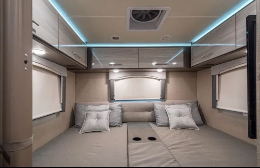 Comfortable RV bedroom interior features a large bed with cup holders, modern overhead storage with blue accent lighting, ideal for an rv rental.. Thor Motor Coach Vegas 2020