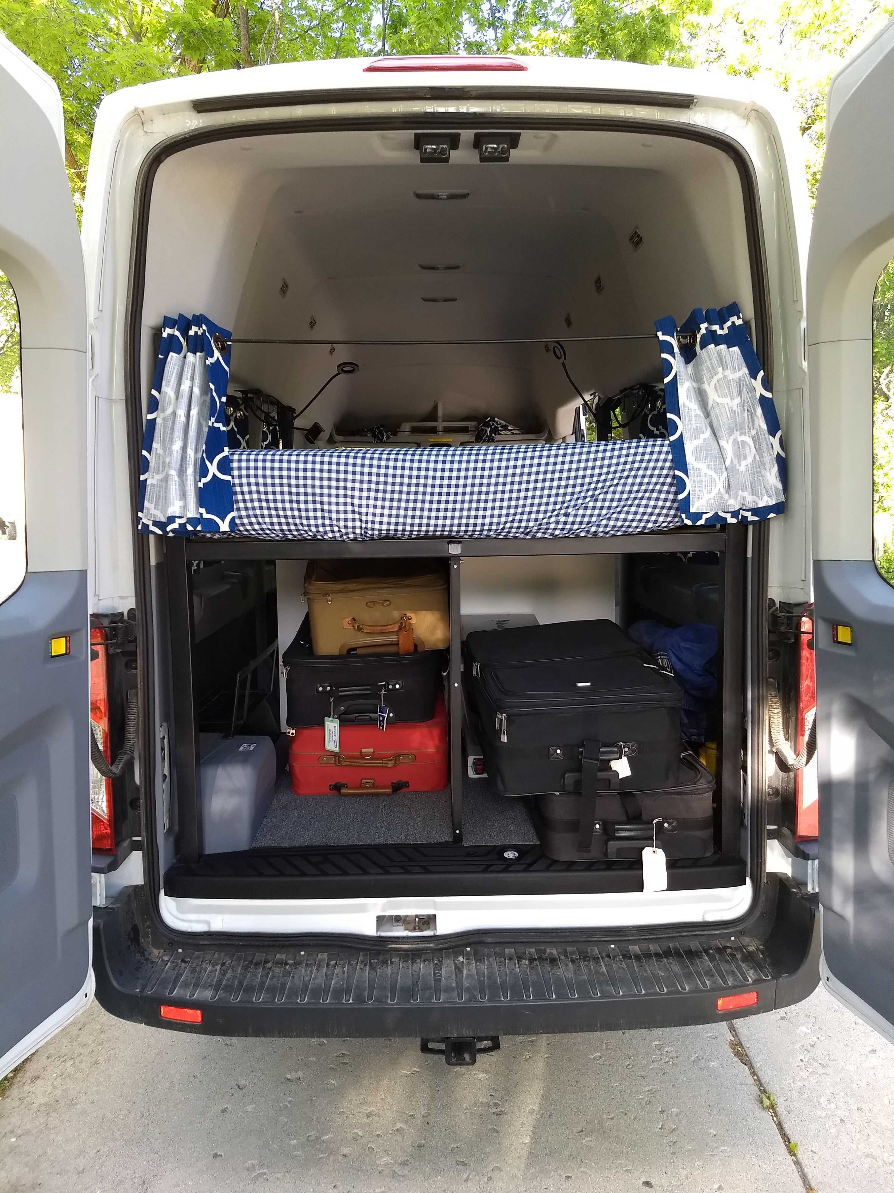 Rear view of an RV rental: organized interior with a raised bed, patterned curtains, and stacked luggage storage.. Ford transit 2020