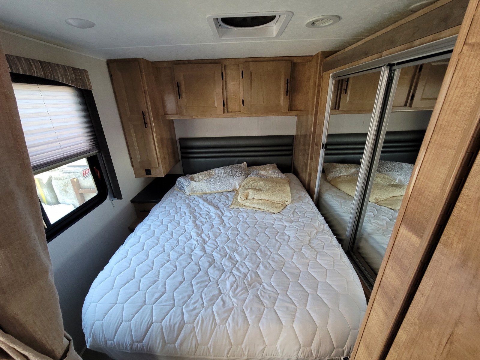 Experience comfort in this RV rental bedroom: bed with white quilt, wood cabinets, and mirrored closet. Ideal for travel.. Coachmen Leprechaun 2021