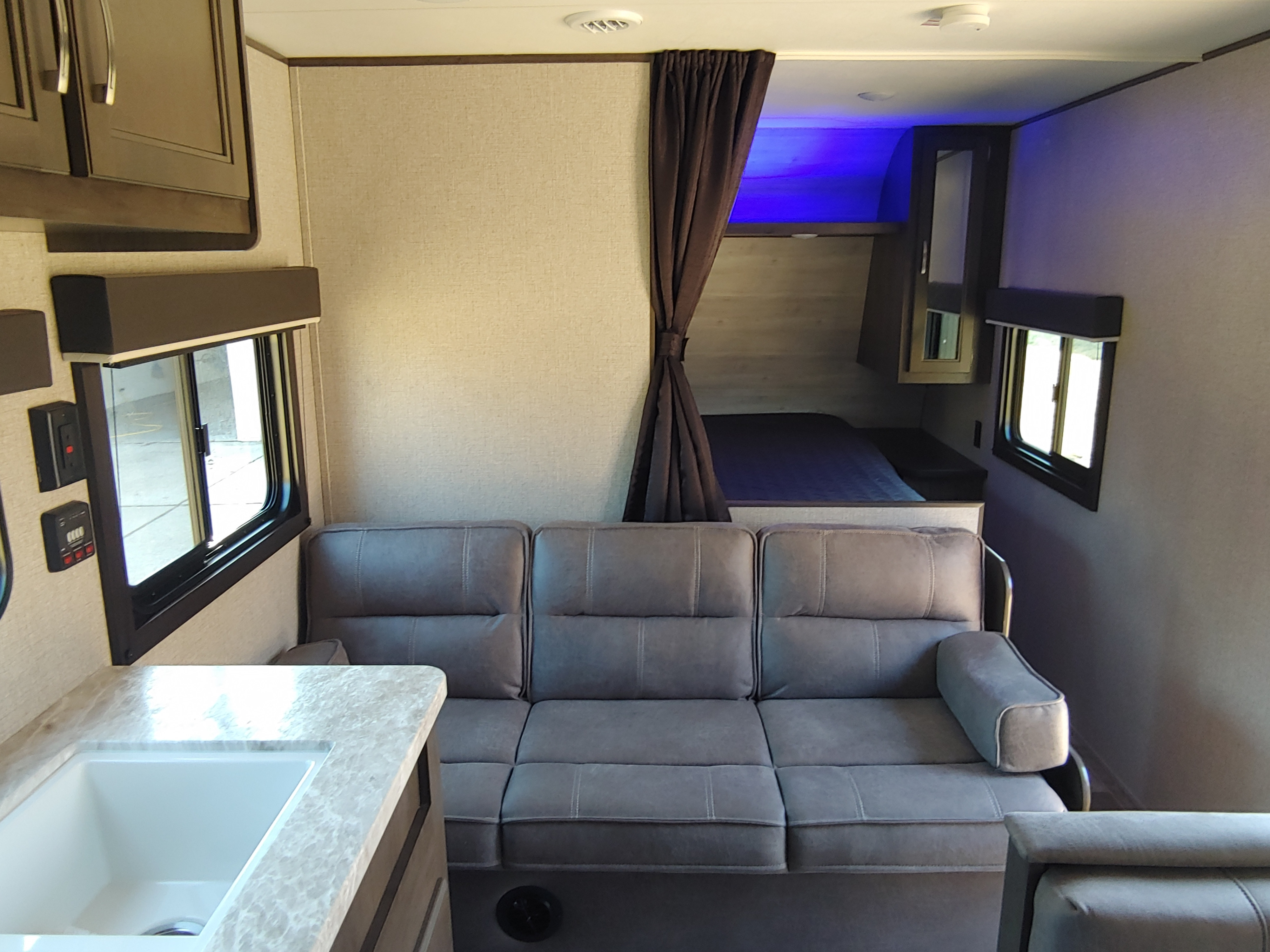 RV rental interior: comfy grey sofa, kitchen sink, and a sleeping area with blue light, separated by a brown curtain.. Jayco Jay Flight SLX 2021