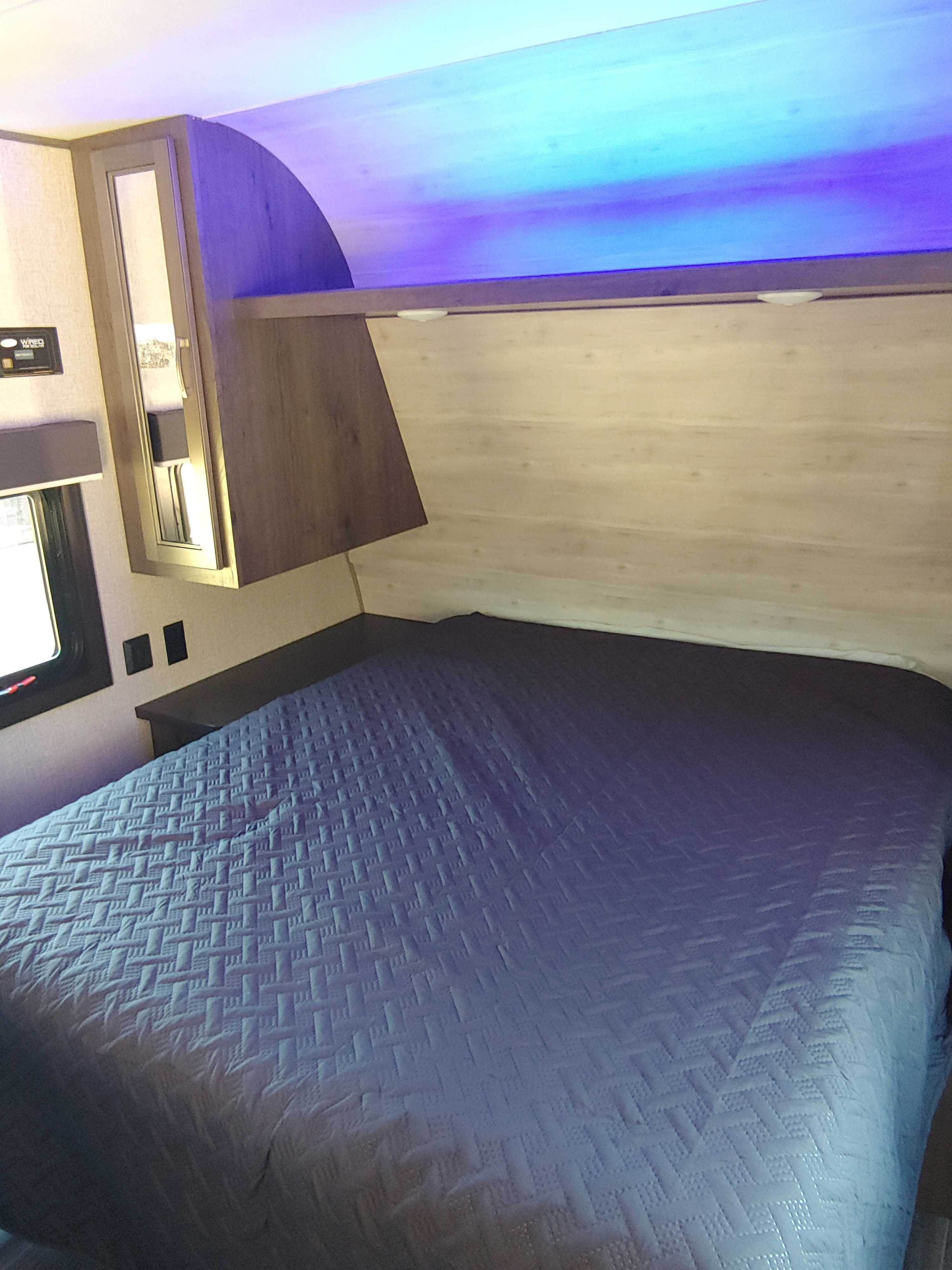 Cozy RV rental bedroom with blue quilted bed and ambient lighting, featuring overhead storage and a window.. Jayco Jay Flight SLX 2021