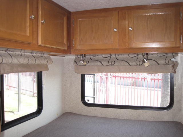 Cozy RV rental interior showing wood cabinets, patterned blinds, and a bed area with reading lights near windows.. Coachmen Leprechaun 2019
