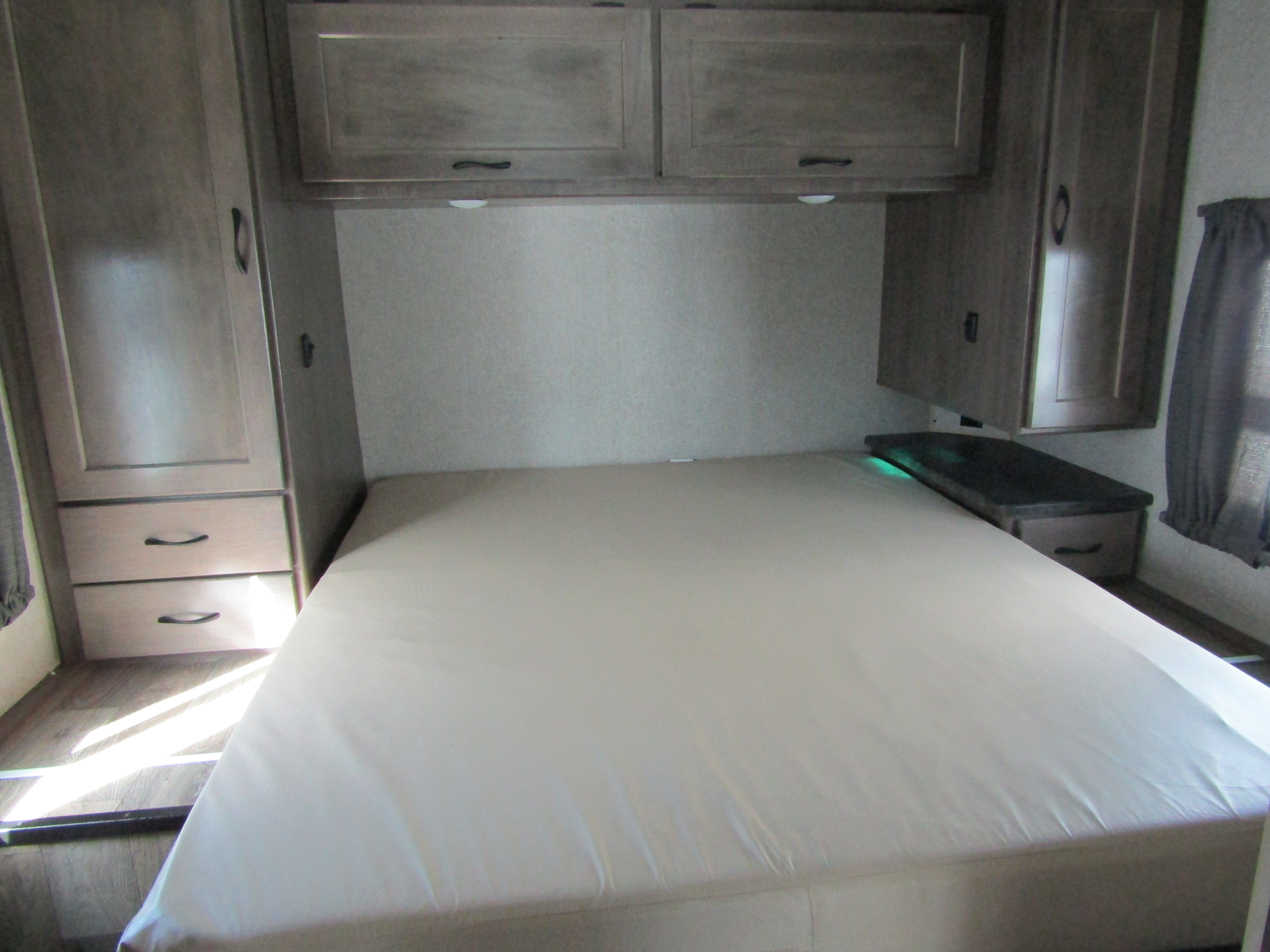 Inside an RV rental, a compact bedroom features a bed, wood-toned overhead cabinets, and a small nightstand with a green light.. Winnebago Minnie Winnie 2020