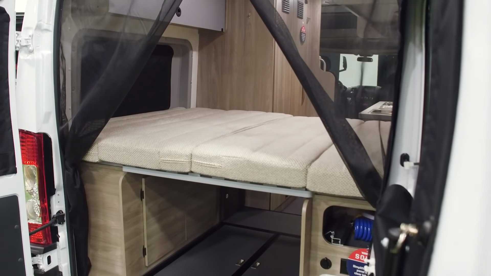 Interior of an RV rental van: convertible bed, under-bed storage, and a black mesh screen at the open rear doors.. Winnebago Solis 2021