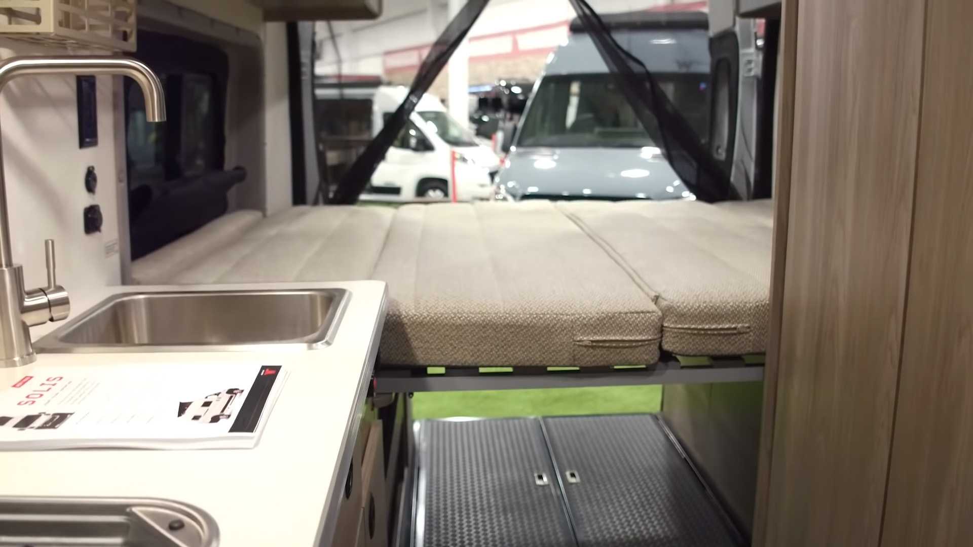 RV rental interior: A camper van's kitchen sink and raised bed are visible. The open rear shows other RVs at a show.. Winnebago Solis 2021