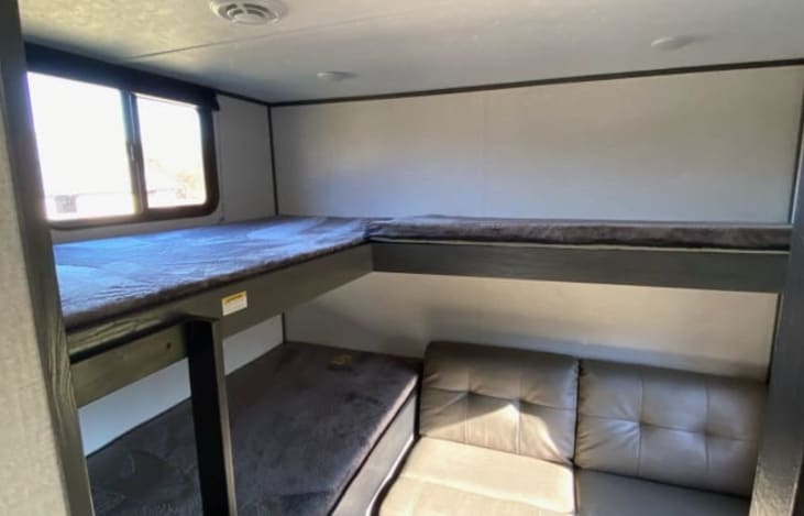 Interior view of an RV rental's bunkhouse. It features multiple stacked beds and a light-colored sofa, with a window on the left.. Aspen Other 2021