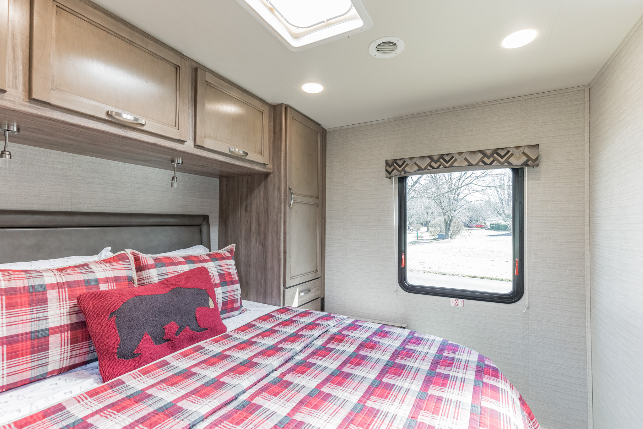 Cozy RV rental bedroom with red plaid bedding, wooden cabinets, and a window displaying an outdoor scene and an 'EXIT' sign.. Jayco Redhawk 2020