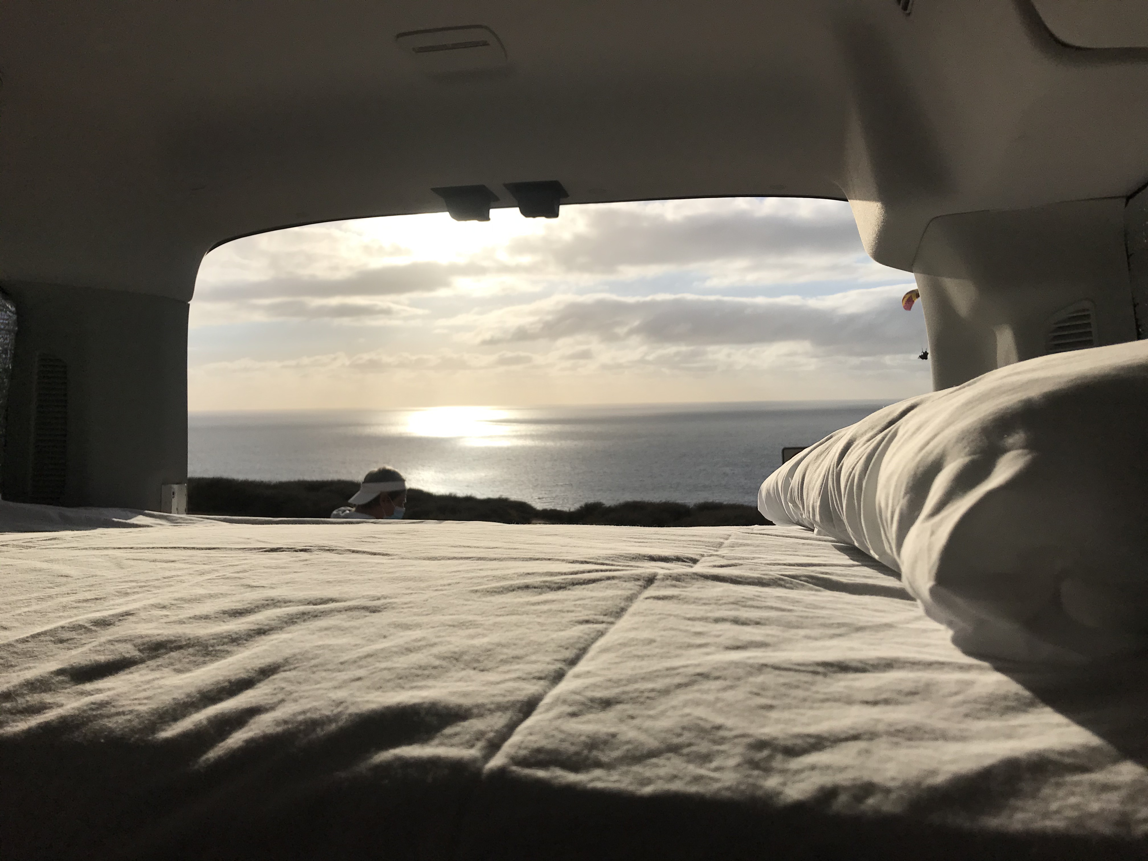 Relaxing with an ocean view from the bed of an RV rental. Sunlight glints on the water; a person watches, and a paraglider flies.. Ford Van 2017