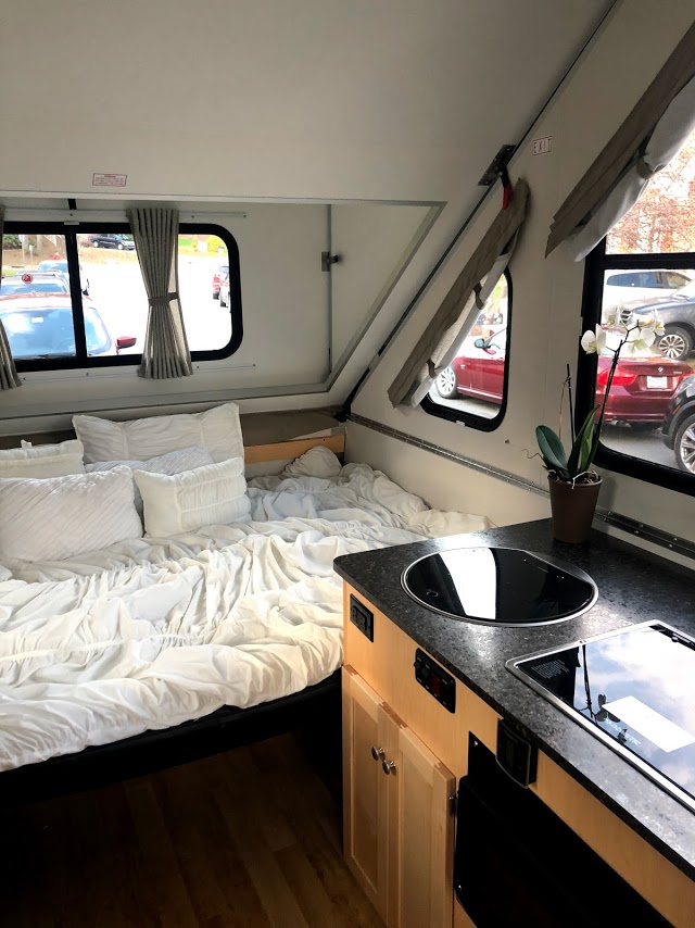 Inside an A-frame RV rental: a cozy bed with white bedding, kitchen counter with sink & stovetop, and natural light from windows.. A-Liner Expedition 2021