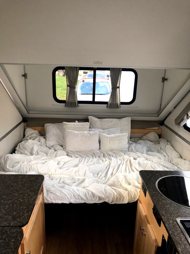 Comfy white bed inside an rv rental, with pillows and windows showing an outside car. A compact travel living space.. A-Liner Expedition 2021