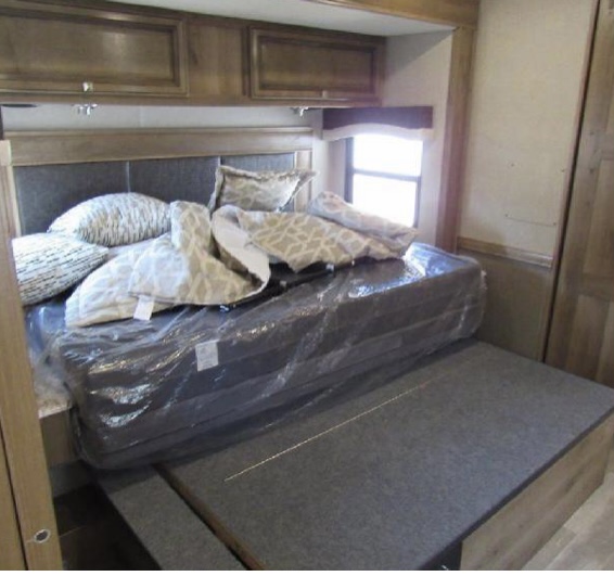 Inside an RV rental bedroom: A compact bed with a new, plastic-wrapped mattress, pillows, and wooden overhead cabinets.. Fleetwood Flair 2020