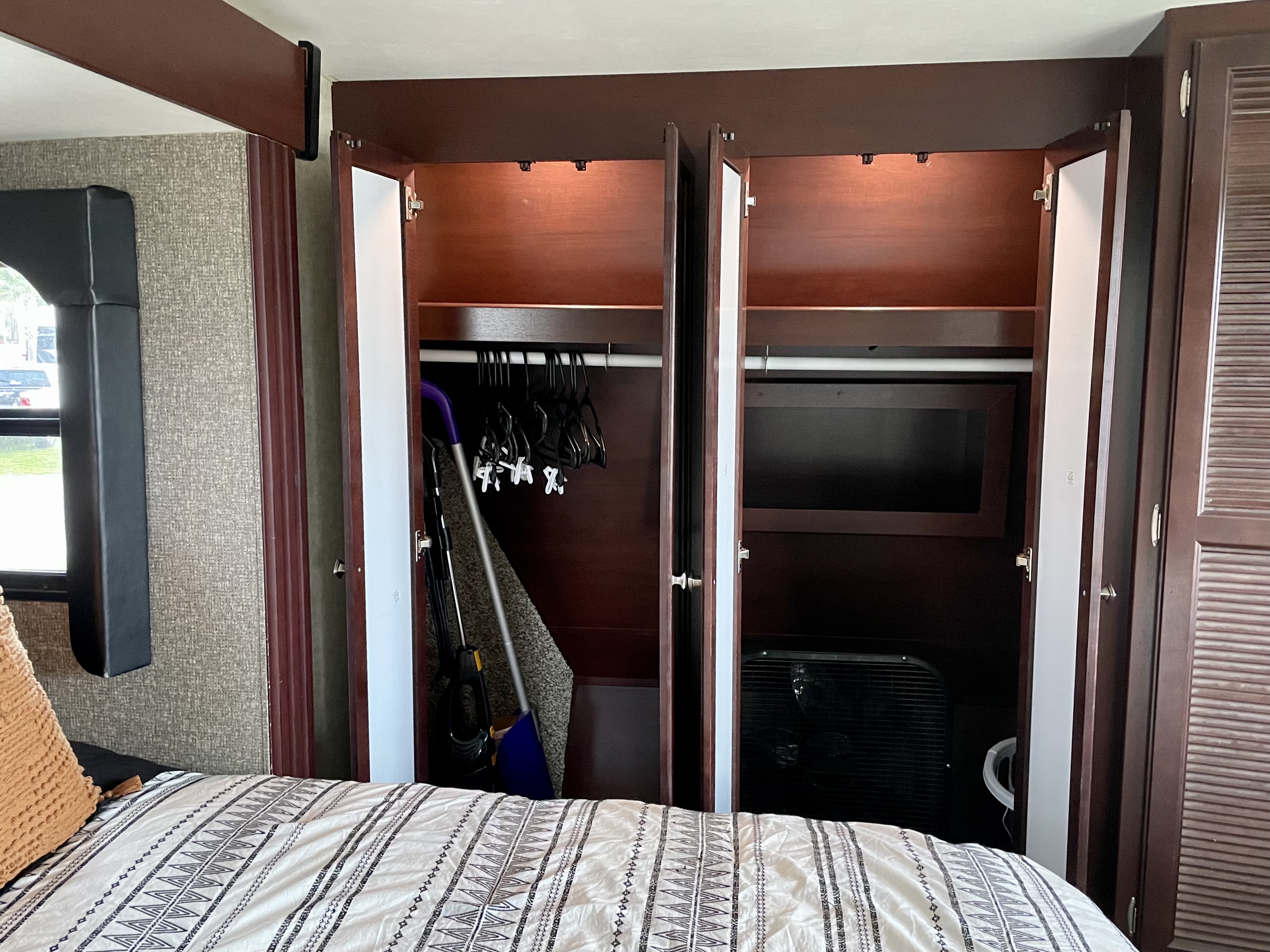 Efficient storage in an RV rental: view inside an open closet with hangers, cleaning tools, and a fan next to a bed.. Heartland RVs Road Warrior Toy Hauler 2015