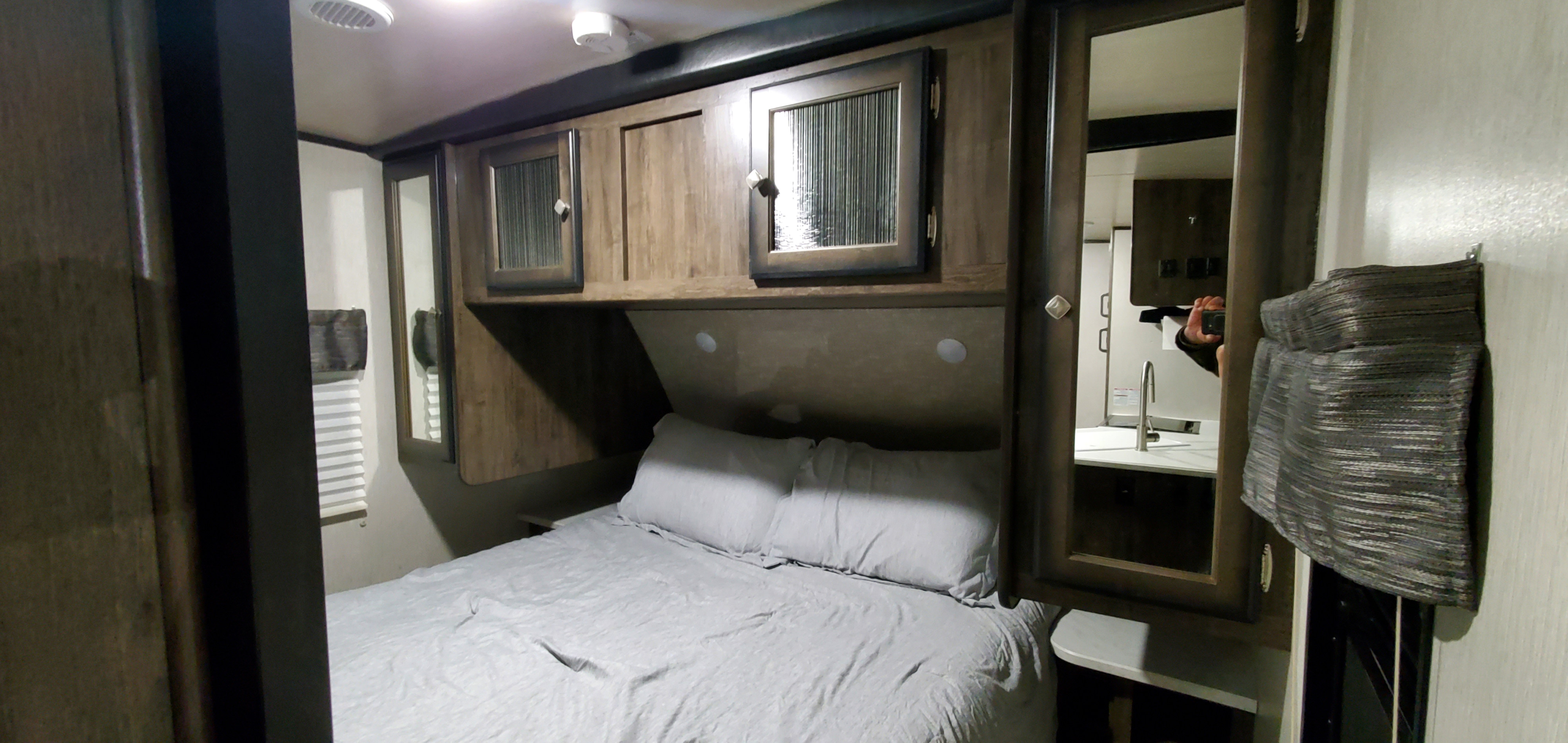 Inside a comfortable RV rental, featuring a queen-size bed with grey linens, wood overhead cabinets, and a reflected kitchen area.. Heartland RVs Mallard 2021
