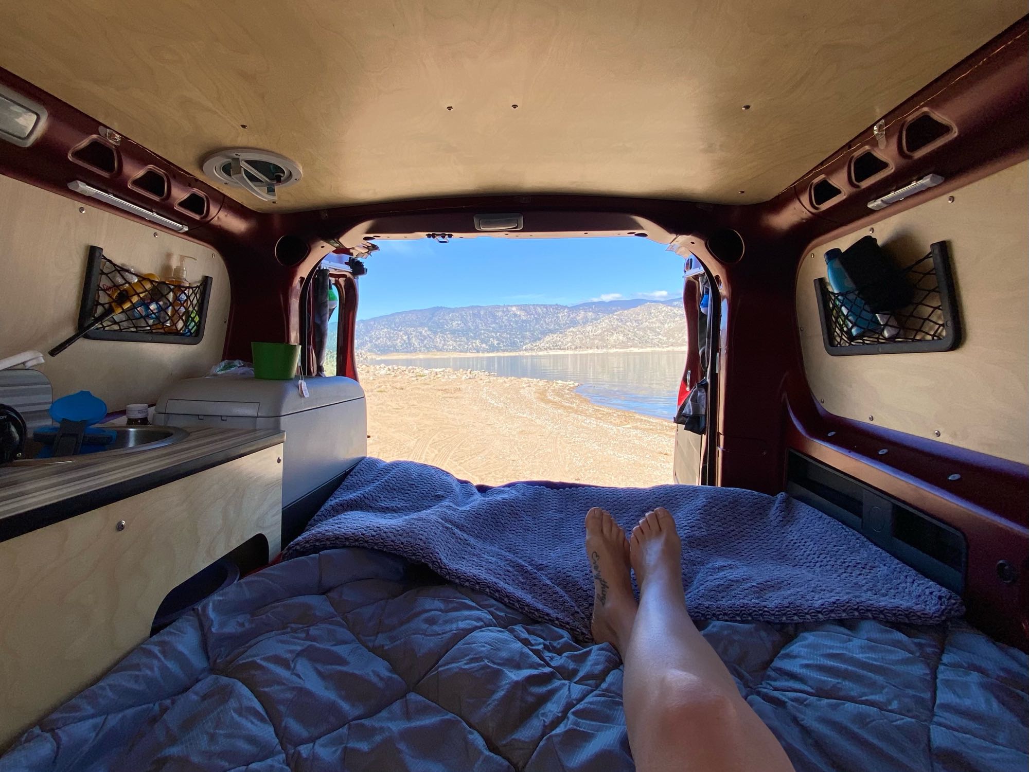 View from inside a camper van: bare feet on a bed, looking out at a lake & mountains. A perfect RV rental escape!. Cascade Campers Ram Promaster City 2018