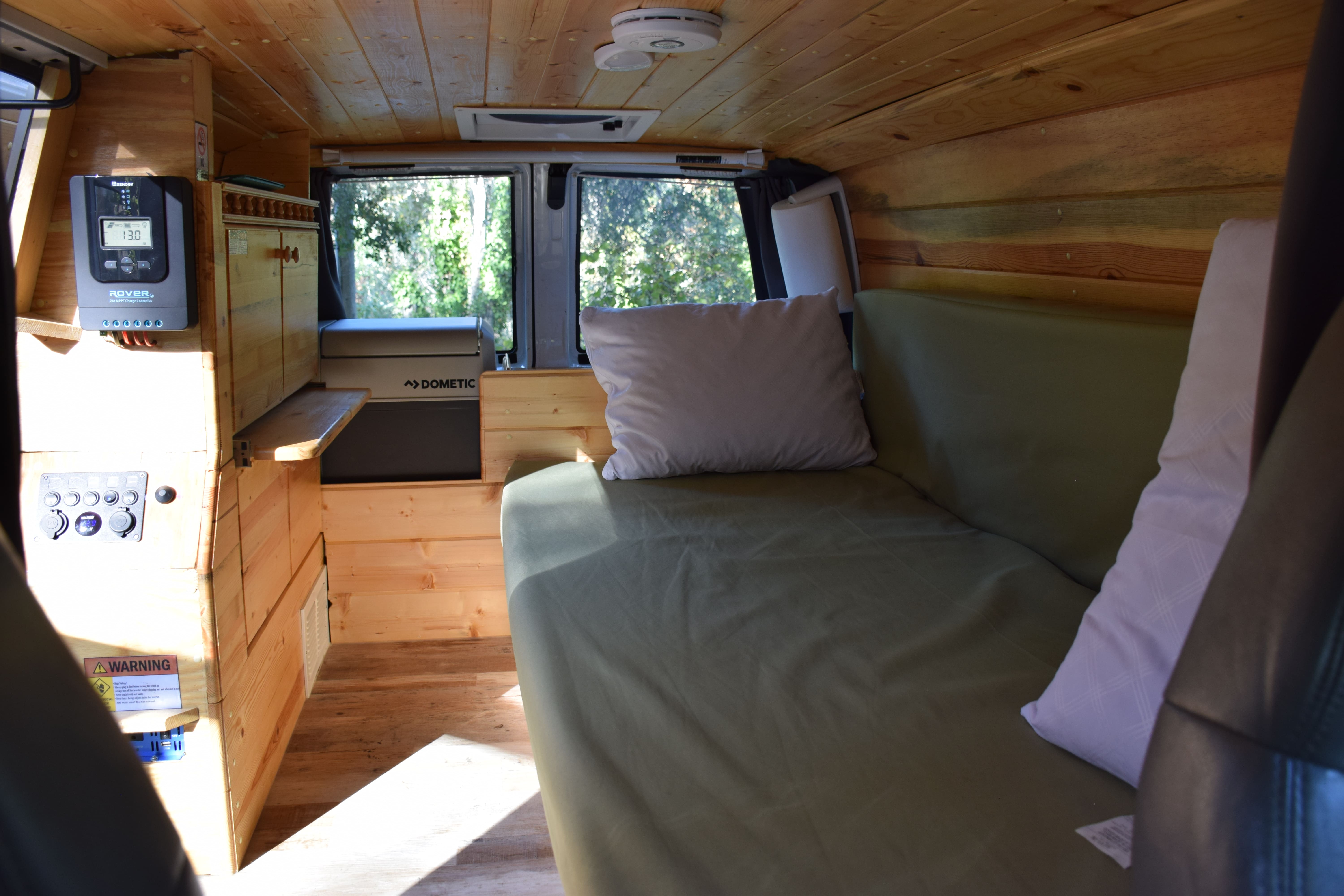 Cozy wood-paneled RV rental interior with a green bench bed, solar controller, and Dometic fridge visible, overlooking trees.. Other Other 2000