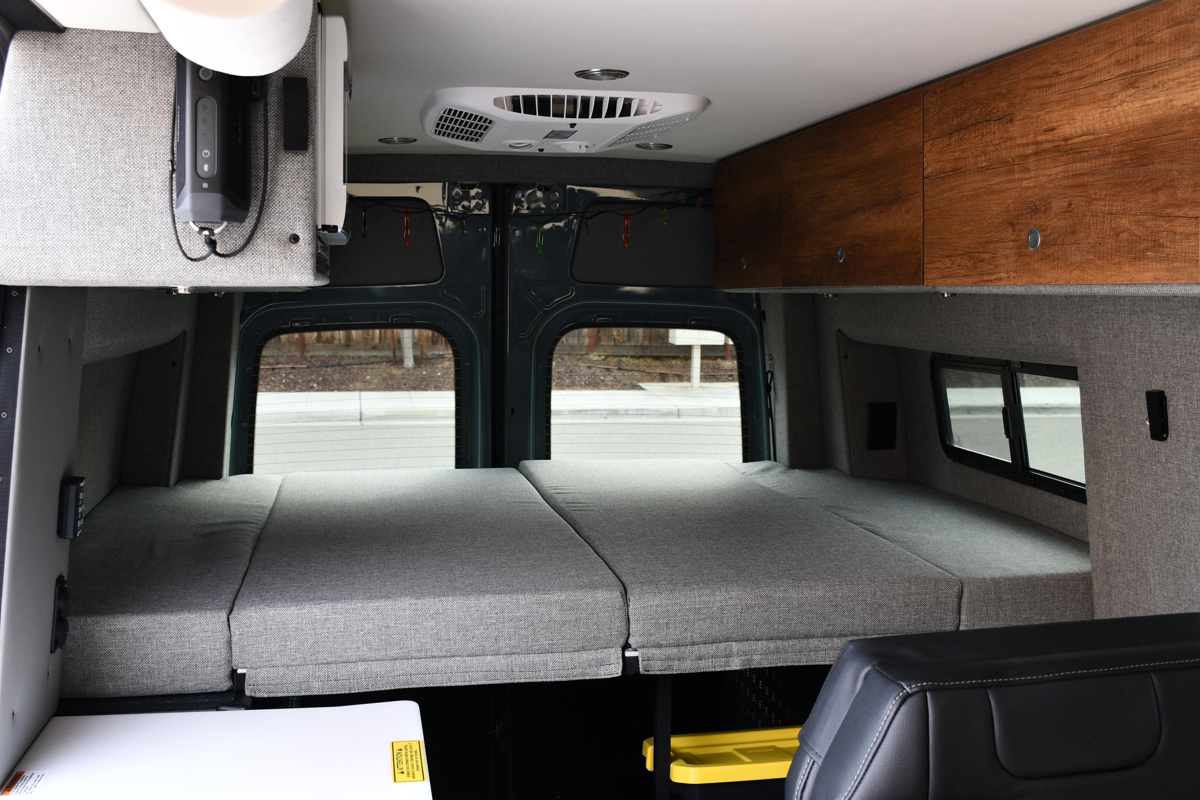 View inside an RV rental's camper van, showing a grey rear bed, wooden overhead cabinets, AC unit, and side windows.. Storyteller Overland Storyteller Overland Class B 2021