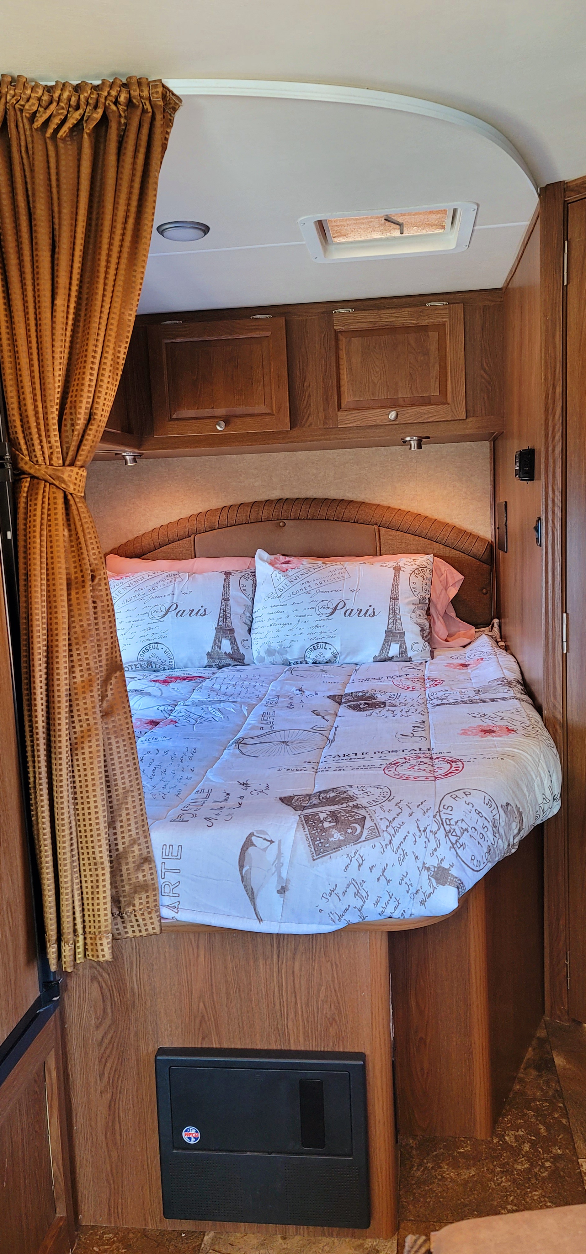 Inside an **RV rental**, a bed with Paris-themed linens, overhead cabinets, and a curtain offers a compact sleeping space.. Coachmen Leprechaun 2016