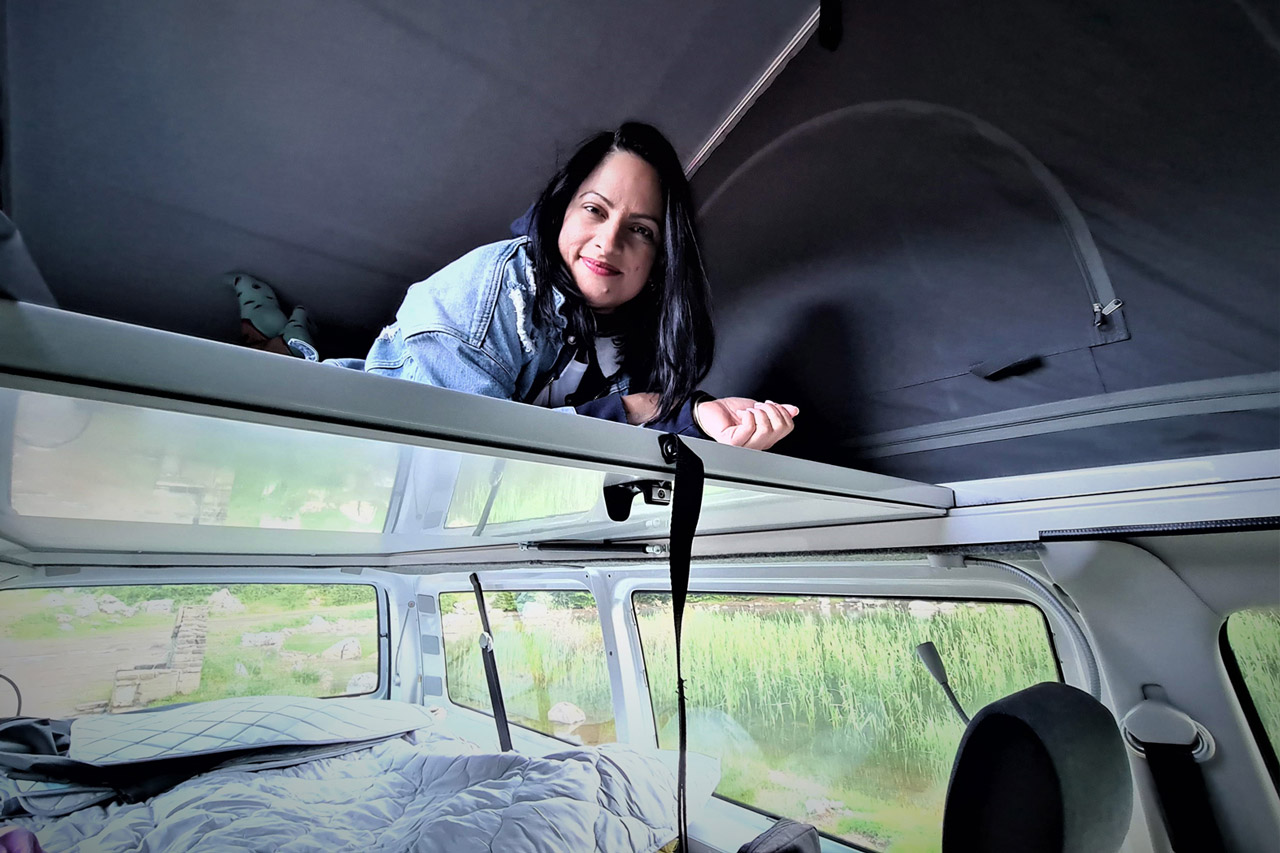 Woman smiles from a pop-top bunk in an RV rental, overlooking a cozy lower bed and green outdoor views.. Volkswagen T6 2020