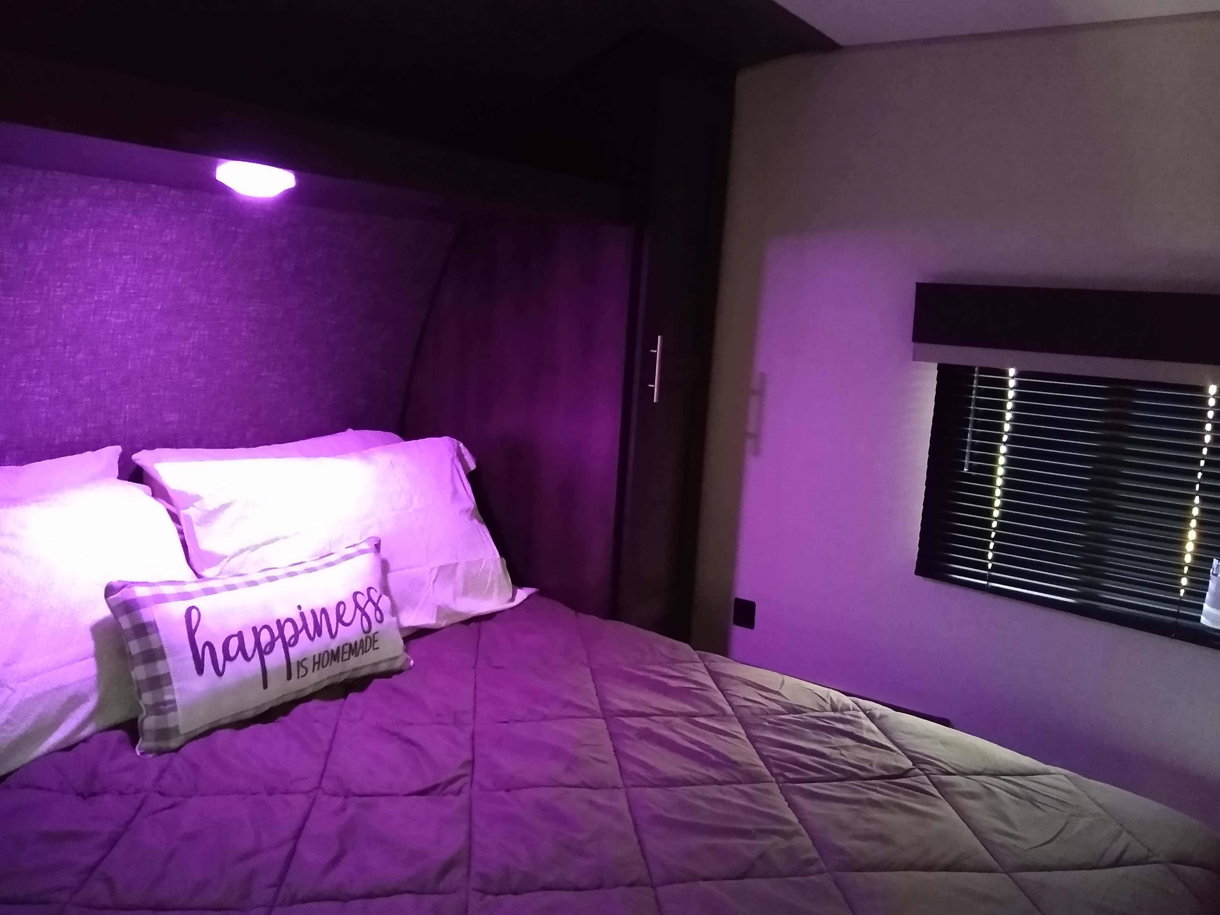 Cozy RV rental bedroom: bed with white pillows, purple comforter, and "happiness is homemade" pillow under purple ambient light.. KZ Sportsmen 2021
