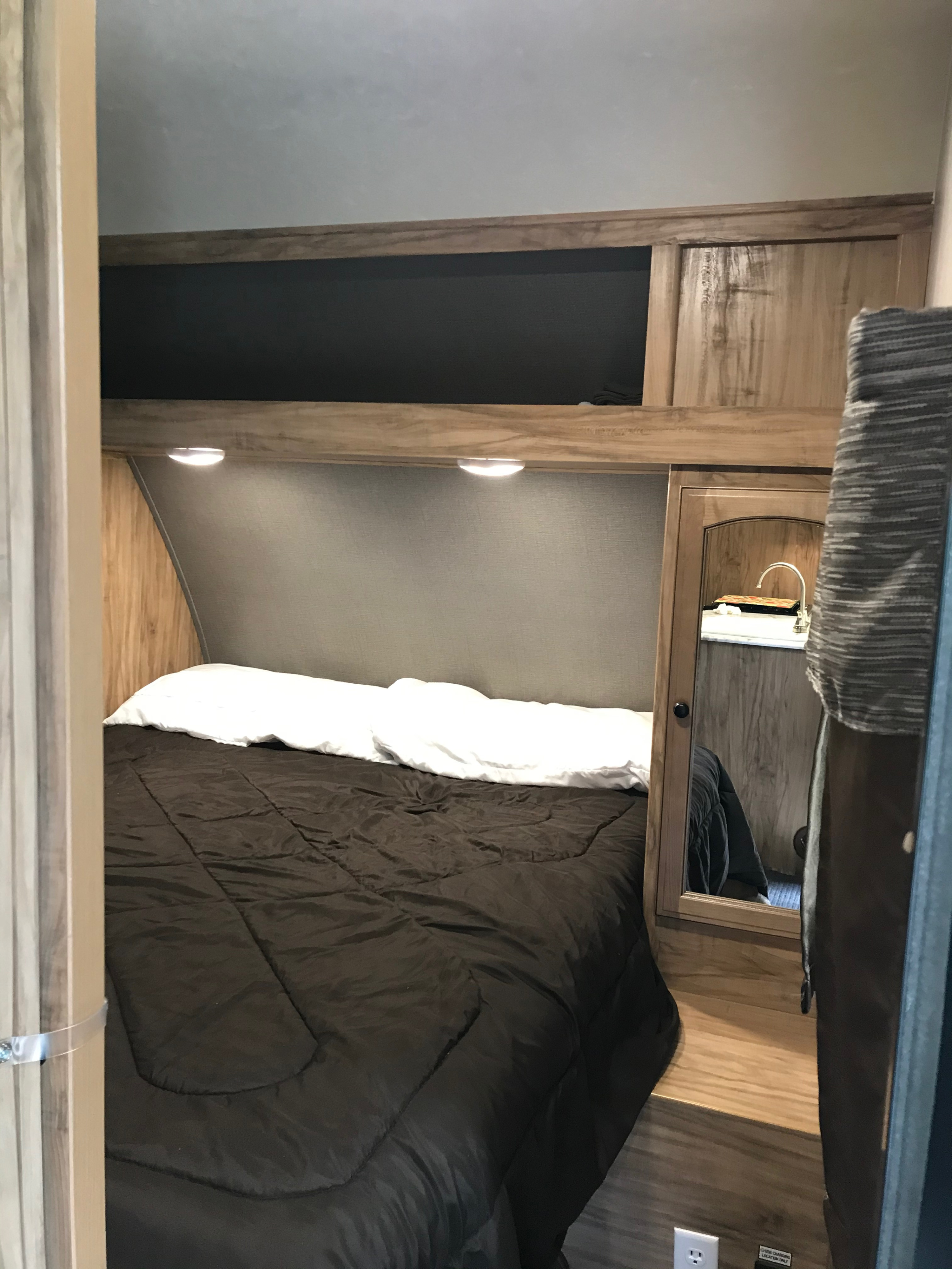 Cozy RV rental bedroom featuring a dark bed, overhead lights, wood cabinetry, and a mirrored sink area.. Coachmen Freedom Express 2018