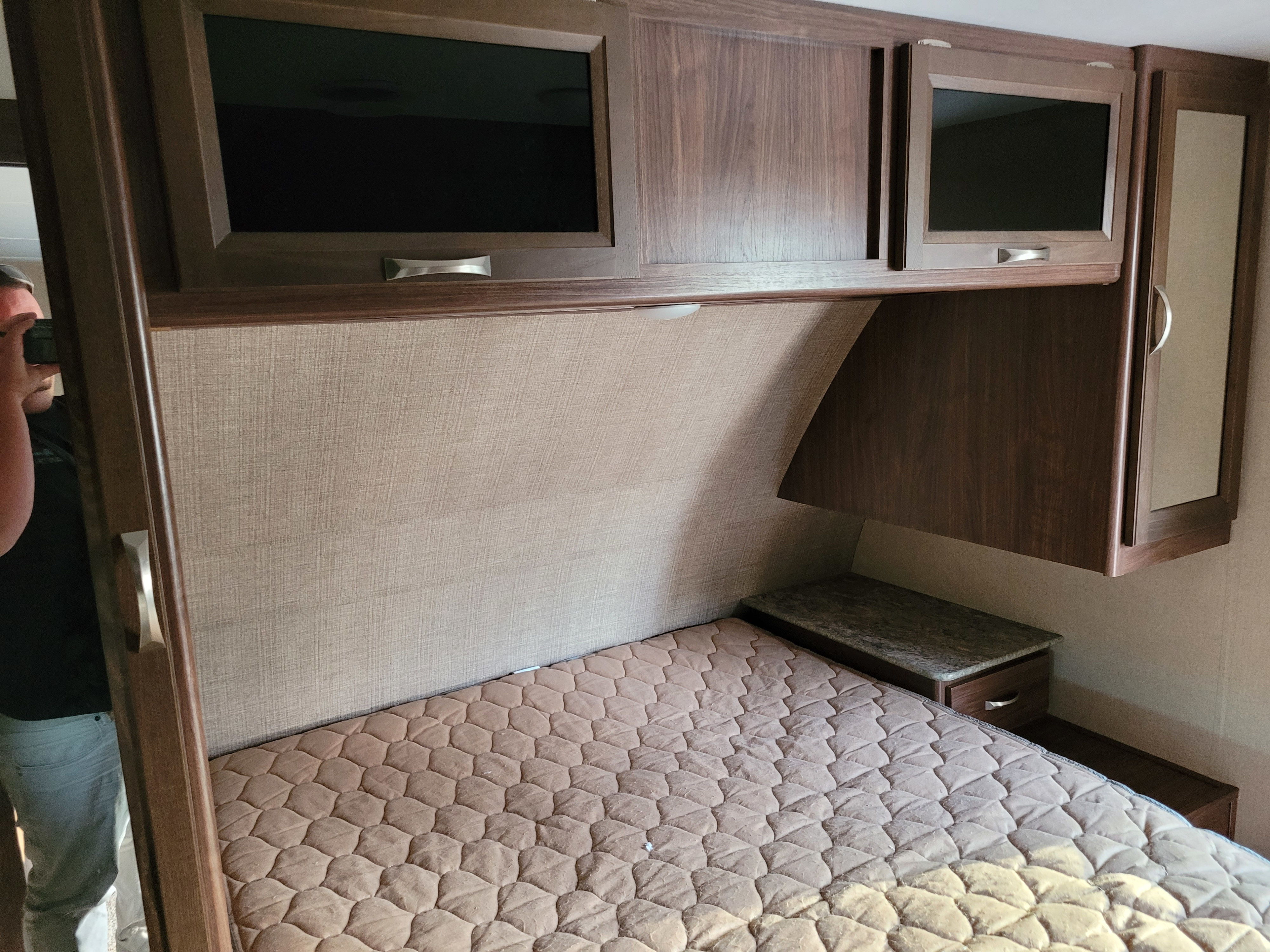 Inside an RV rental bedroom: quilted bed, dark wood overhead cabinets (one with a TV), and a small nightstand, offering compact comfort.. Dutchmen Coleman 2017