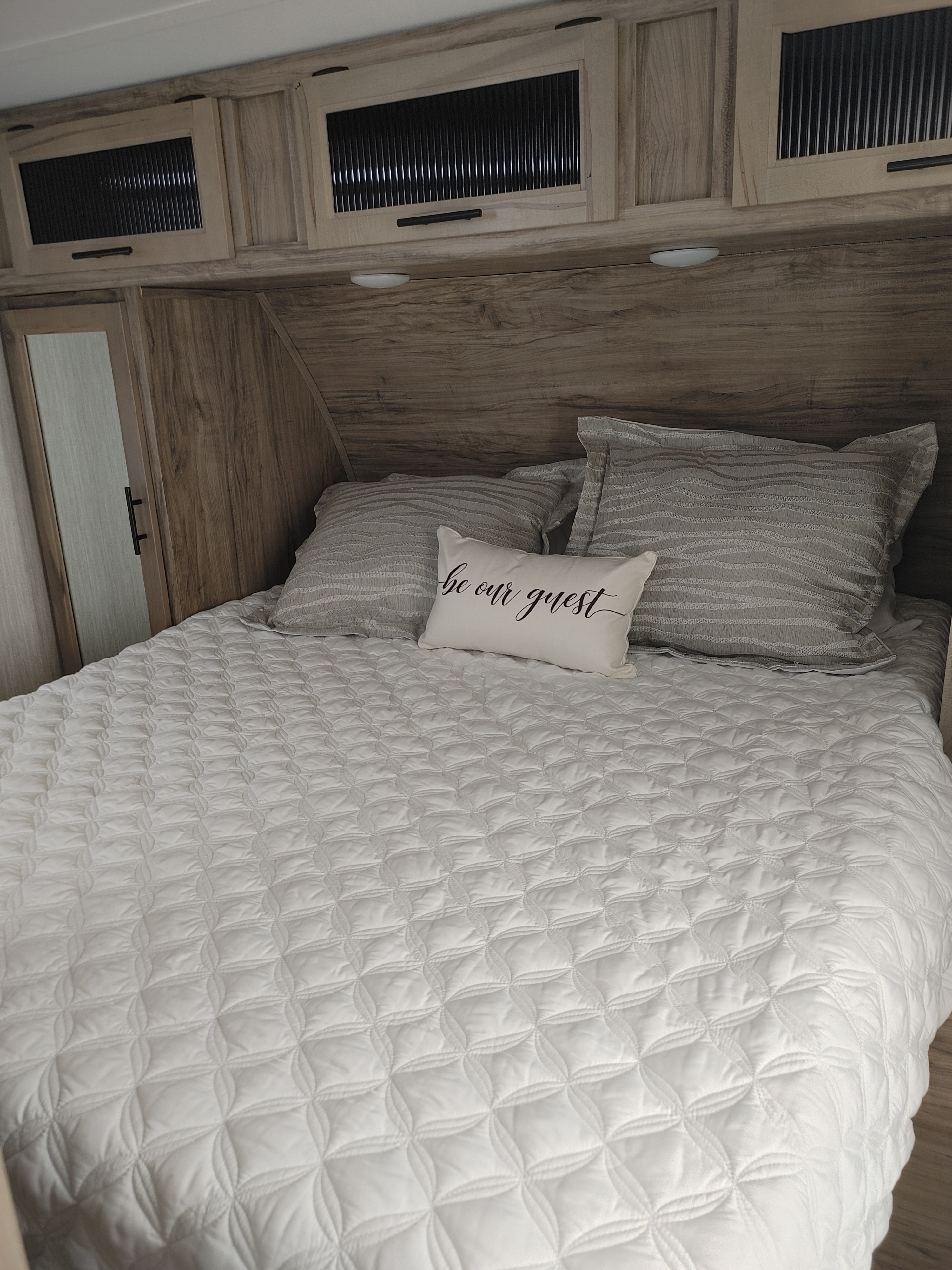 Cozy RV rental bedroom with white quilted bed, gray pillows, and "be our guest" accent pillow, plus wood cabinets.. Forest River Cherokee Alpha Wolf 2021