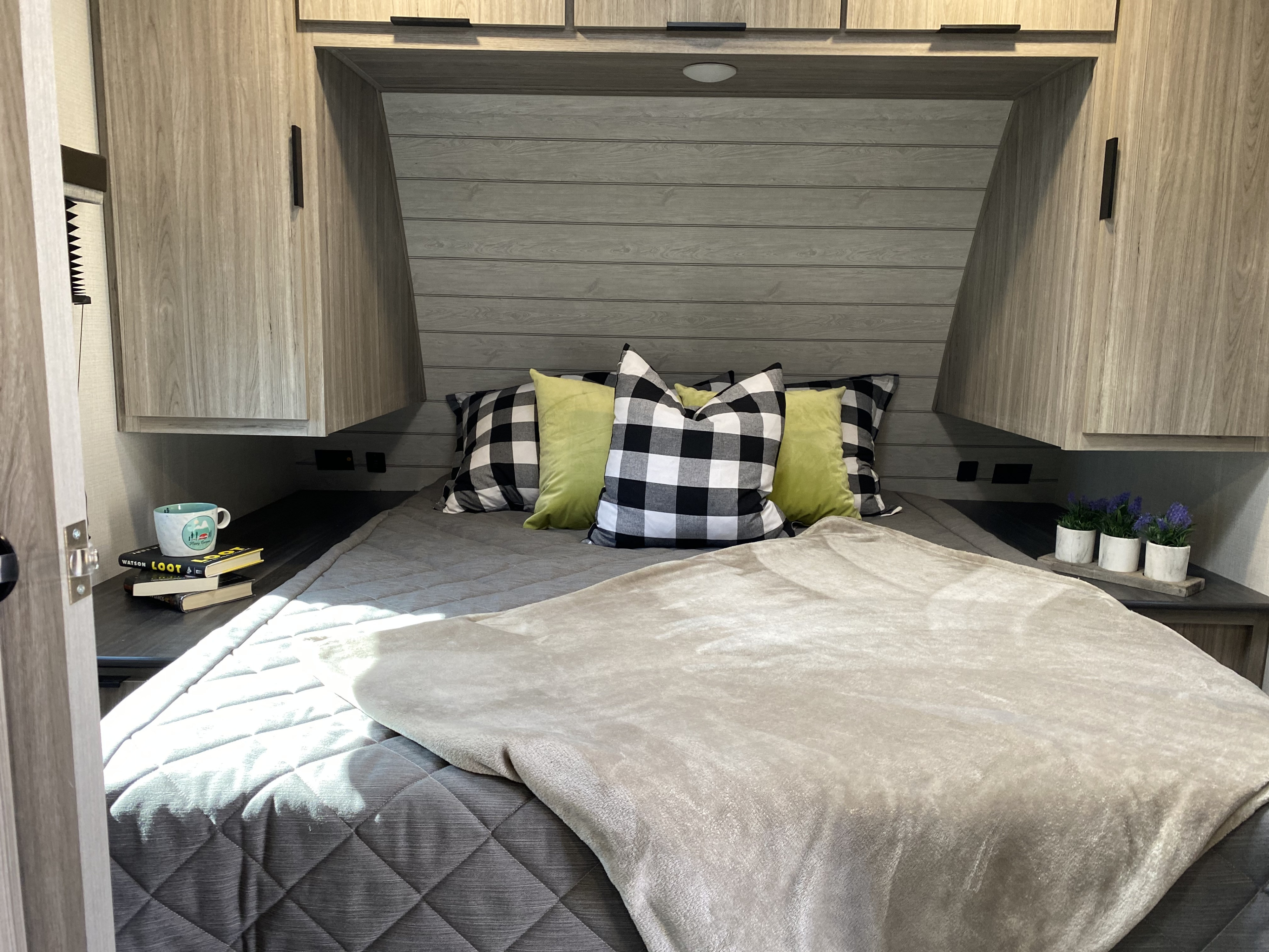 A cozy RV rental bedroom features a gray bed, buffalo check pillows, and light wood cabinetry, ideal for travel comfort.. Keystone Passport 2021
