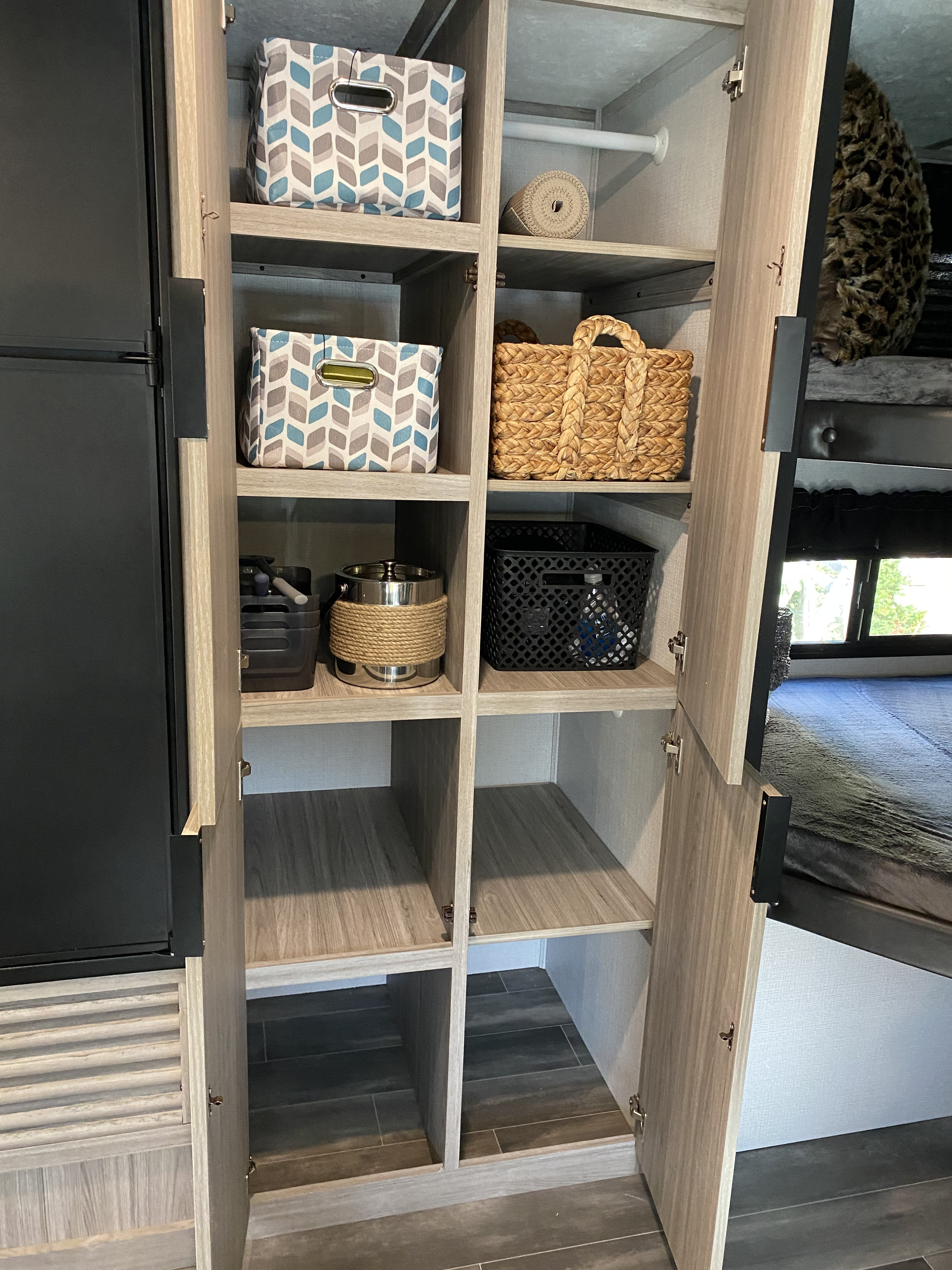 Organized RV rental interior. Open modular shelving with fabric bins, wicker baskets, and other storage next to a fridge and bunk.. Keystone Passport 2021