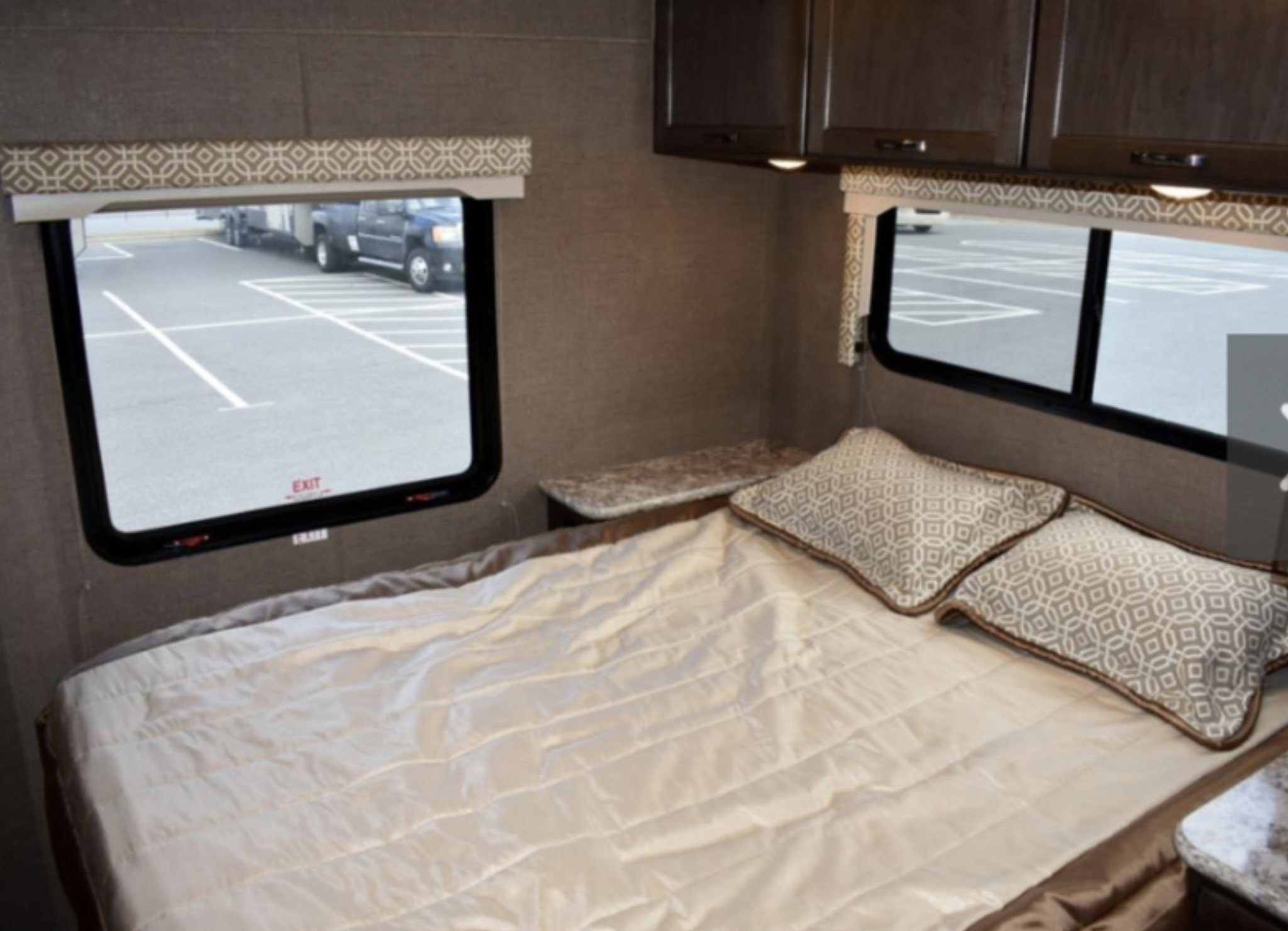 This RV rental bedroom interior features a comfy bed, windows with a parking lot view, and overhead storage cabinets.. Thor Motor Coach Freedom Elite F32 2018