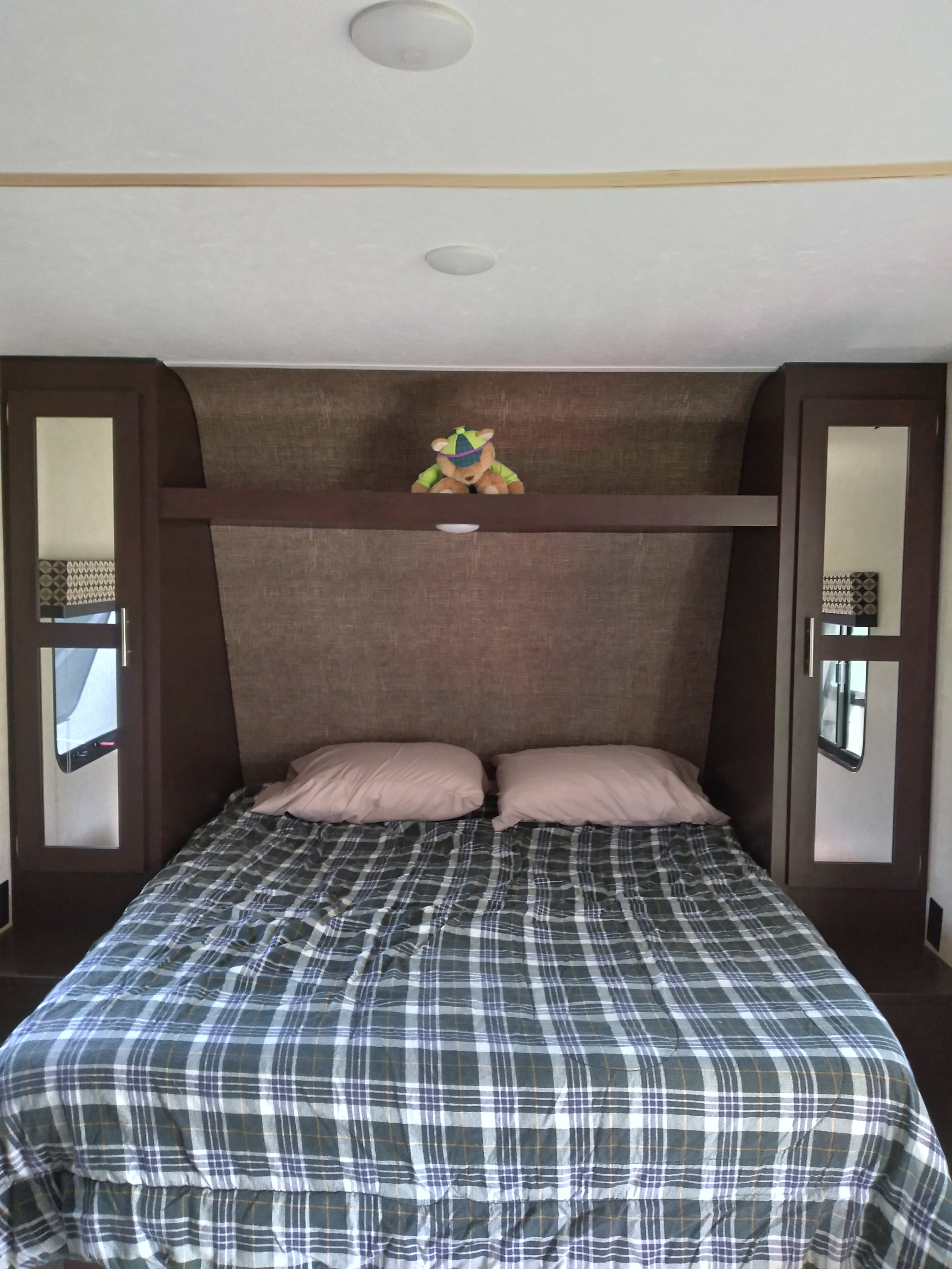 Cozy RV rental bedroom with a plaid bed, pink pillows, and built-in dark wood cabinets. A stuffed animal rests on the shelf.. KZ Other 2017