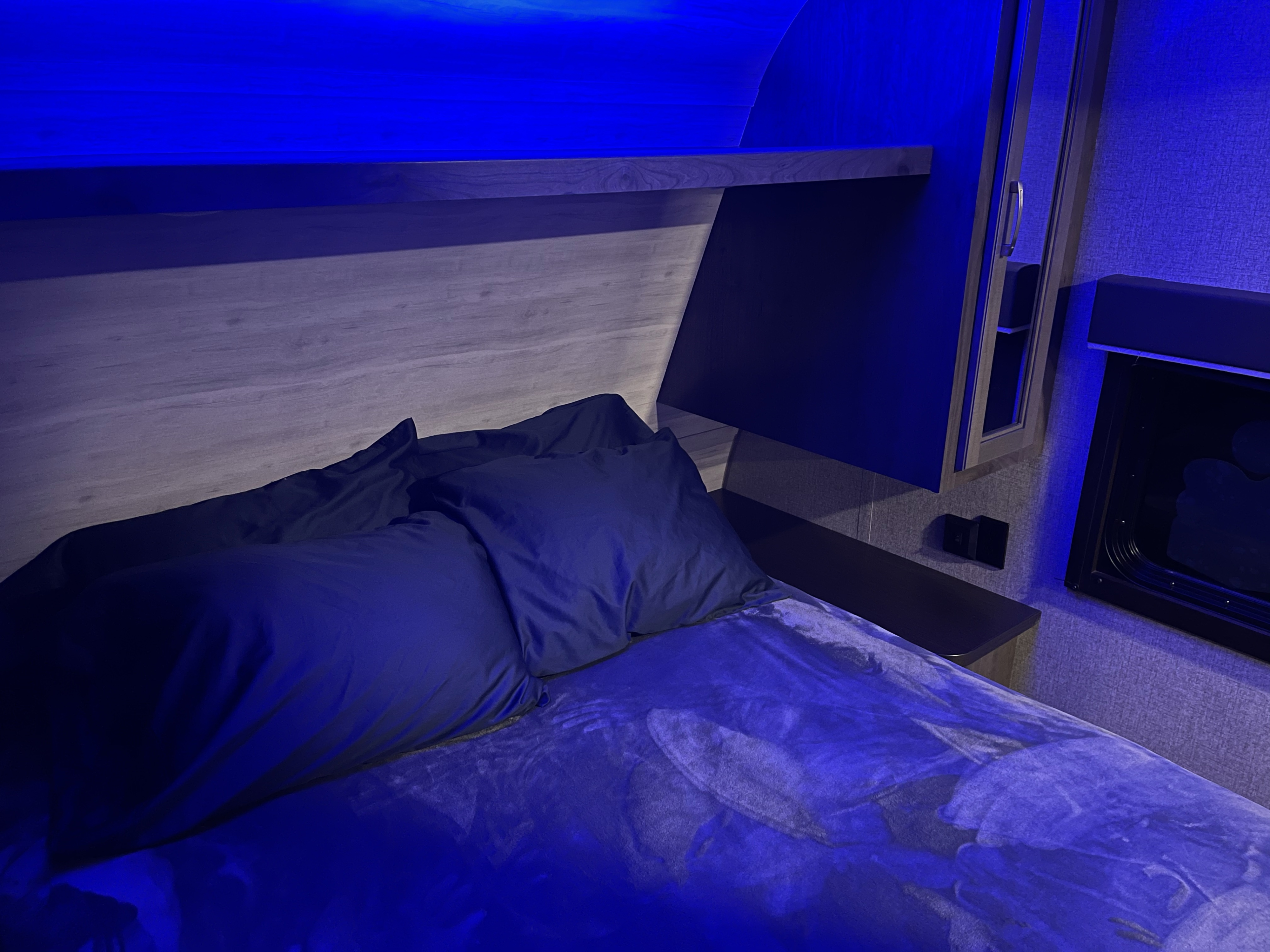 Inside an RV rental, a blue-lit bedroom features dark bedding, a wood-grain headboard, and bedside storage.. Jayco Jay Flight 2021