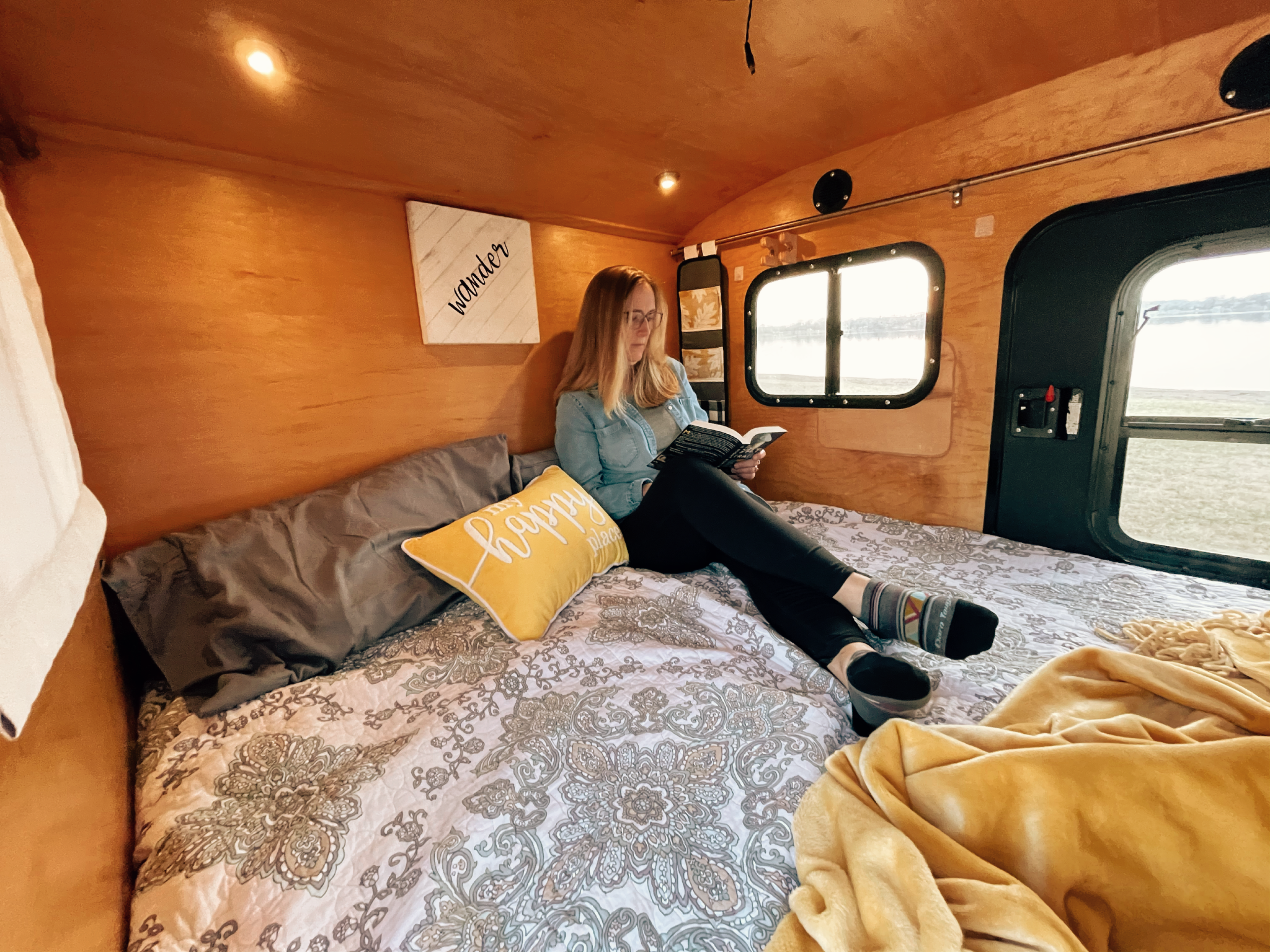 Enjoy a peaceful moment in your next RV rental. A woman reads on the comfortable bed inside a rustic camper with a scenic view outside the window.. American Teardrop Travel Trailer 2016