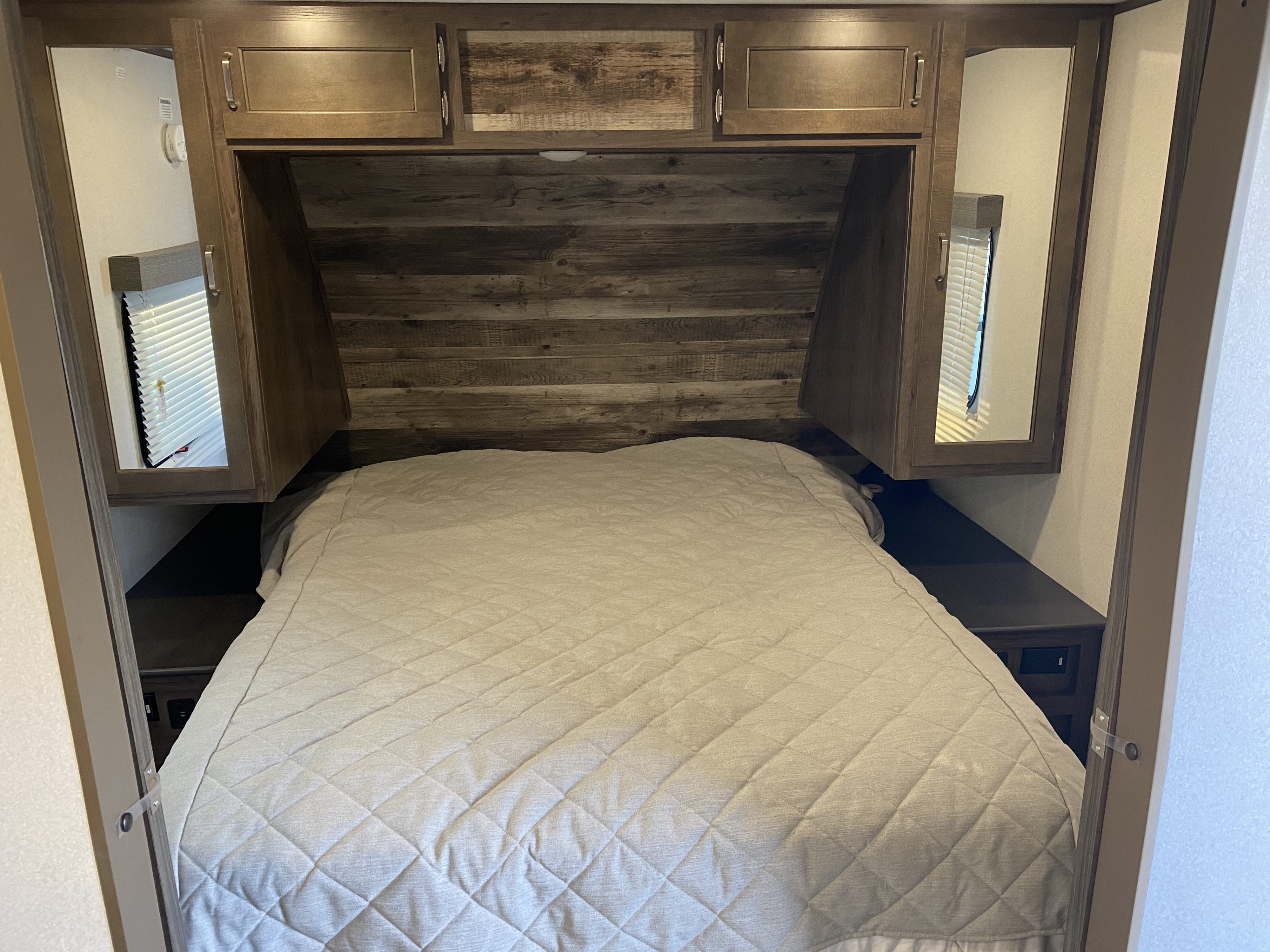Inside this RV rental, a compact bedroom offers a comfy bed, wood headboard, overhead cabinets, and side windows with blinds.. Keystone Passport 2020