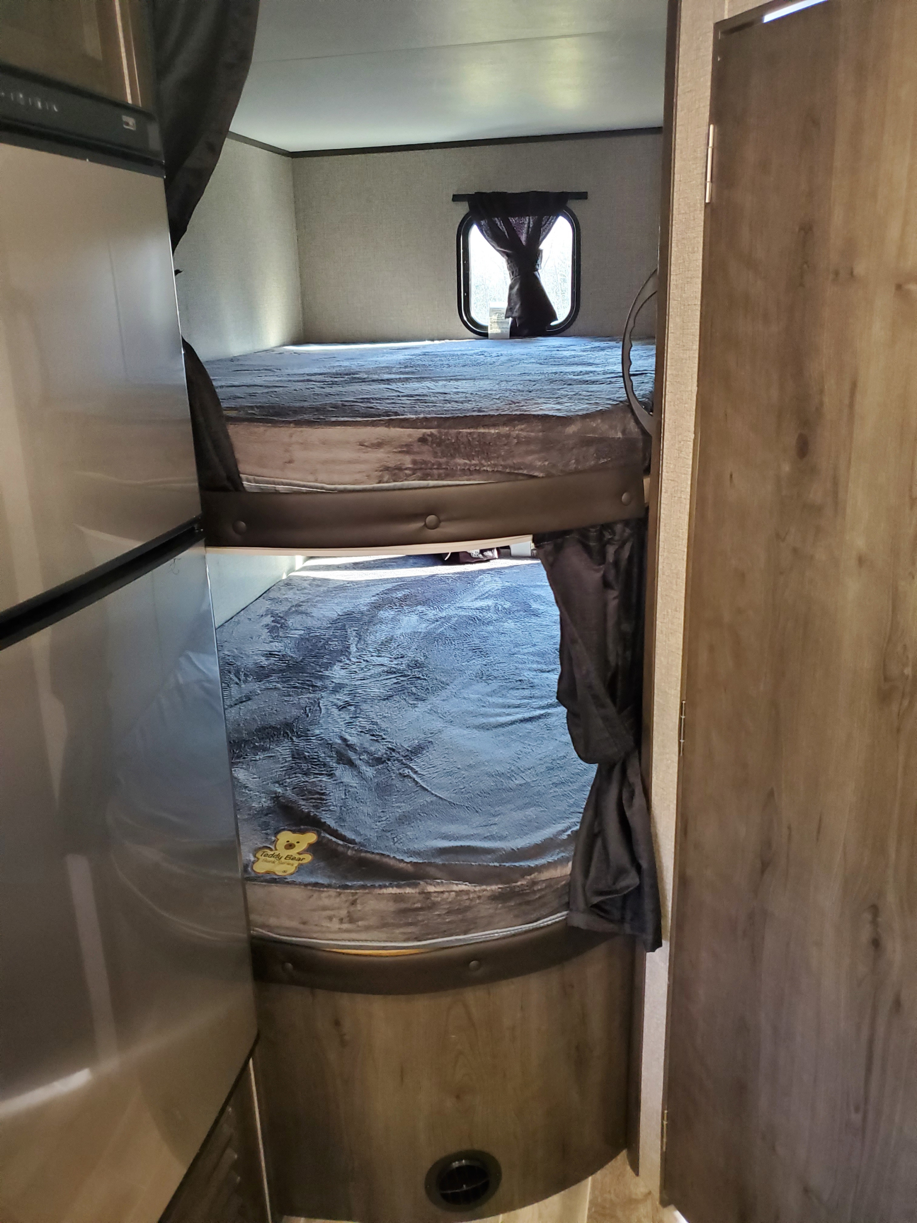 An RV rental interior featuring stacked bunk beds with blue blankets, a window with curtains, and a fridge on the left.. Jayco Jay Flight 2021